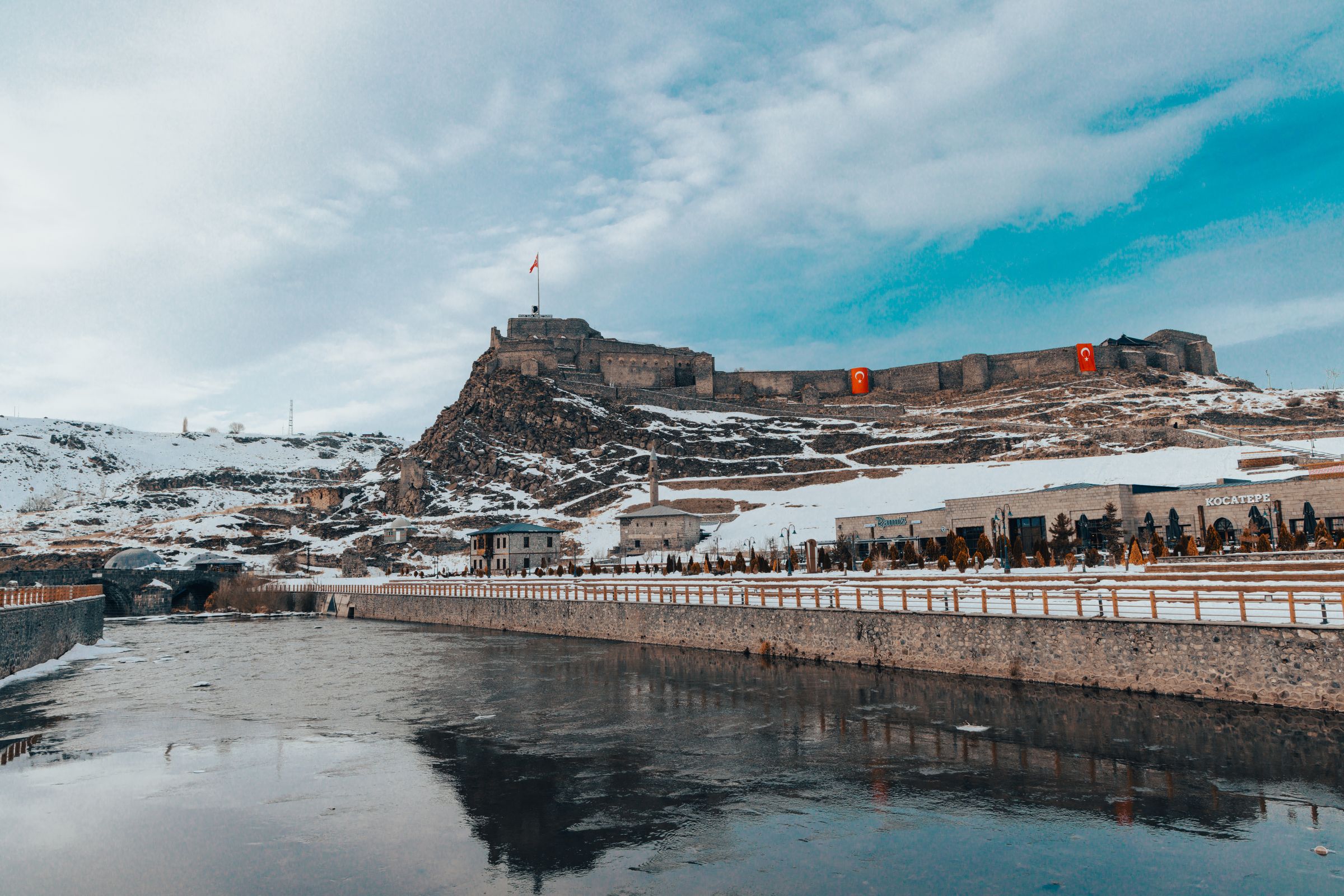 Kars castle on its snowy hill above the river in winter, Turkish flags on the walls and the embankment in the foreground