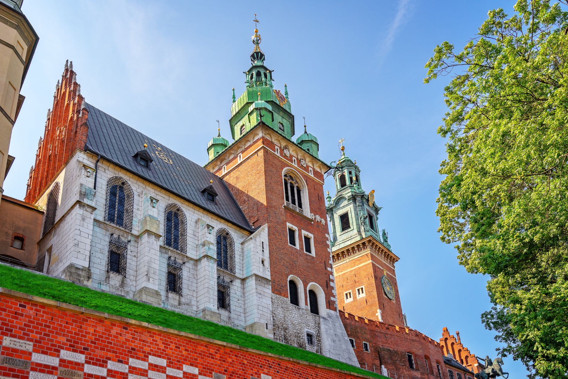 Wawel Cathedral in Krakow, red brick walls and green copper domes over the chapel roofs, seen from below through tree branches