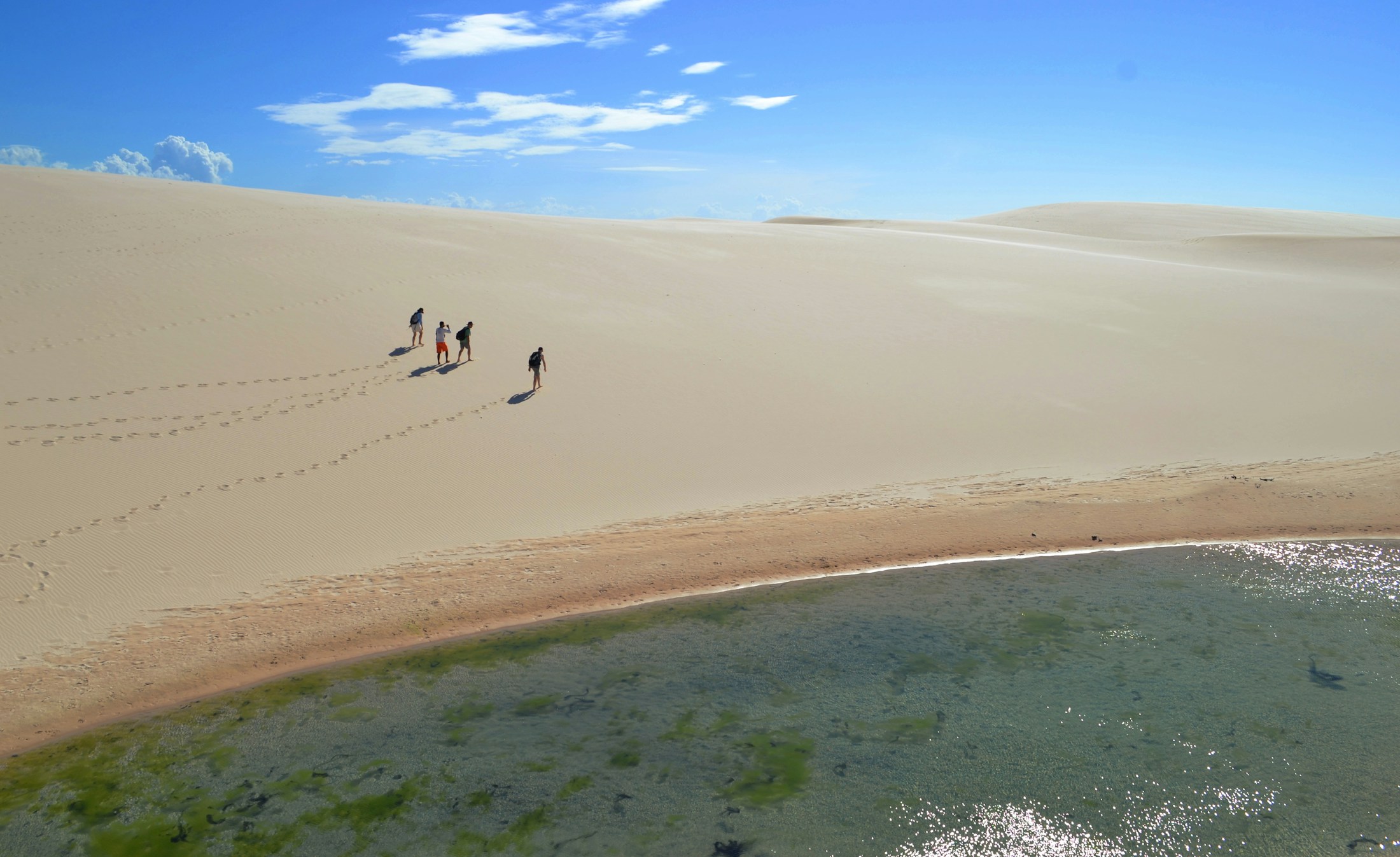A small group of people crossing a vast white dune on foot down toward a lagoon