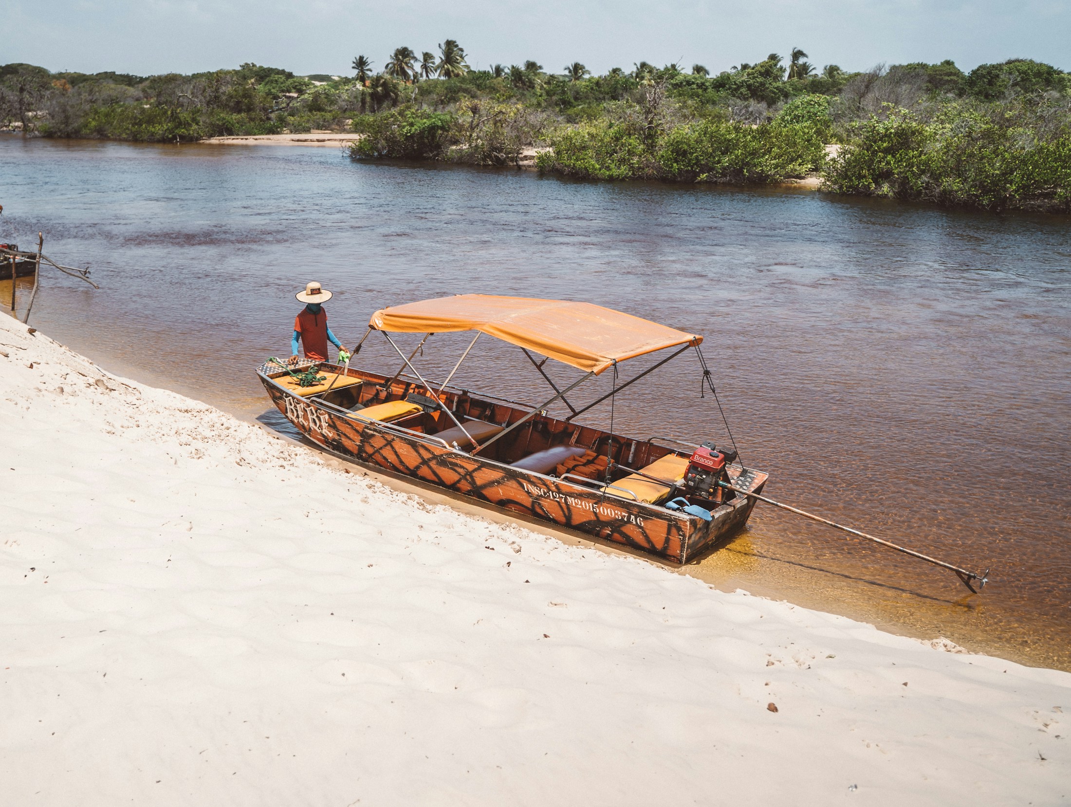 An open wooden boat with an orange canopy on the brown Preguiças river, green banks behind