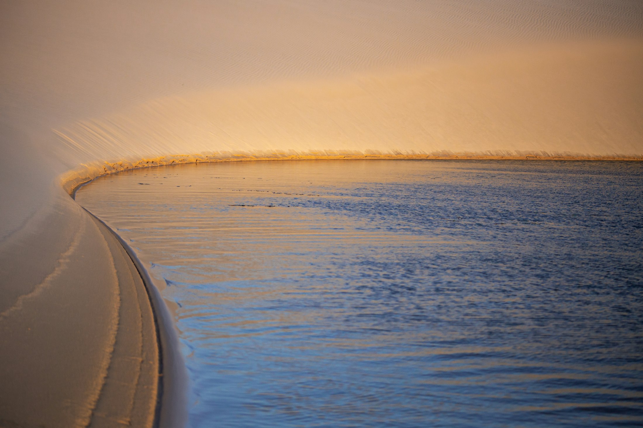 Sunset over the dune field, the sand turning gold and rose, a lagoon holding the colour of the sky