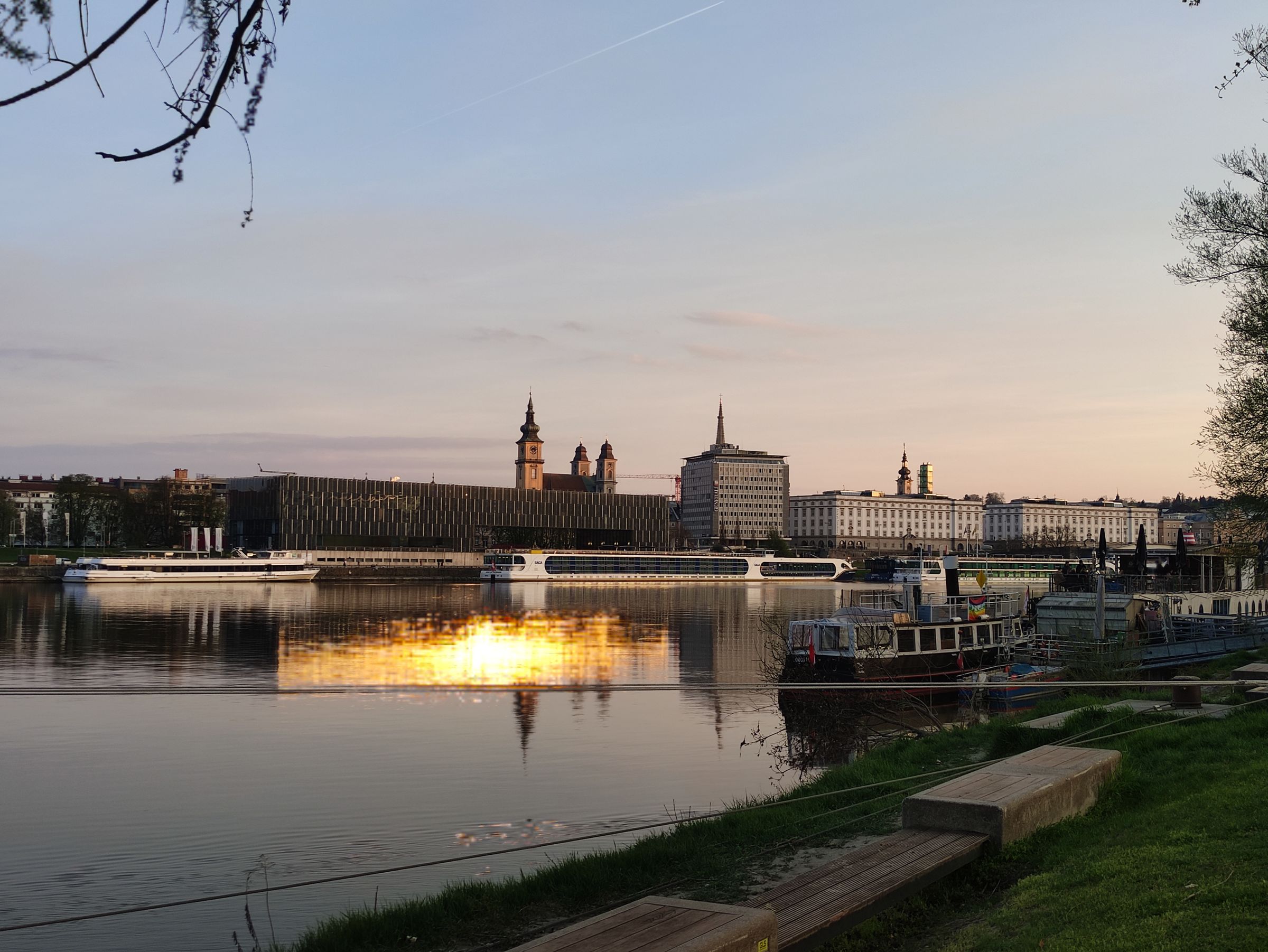 The Danube at Linz at sunset from the north bank, river cruise boats moored below a long dark modern building, church towers and a row of pale buildings on the far side