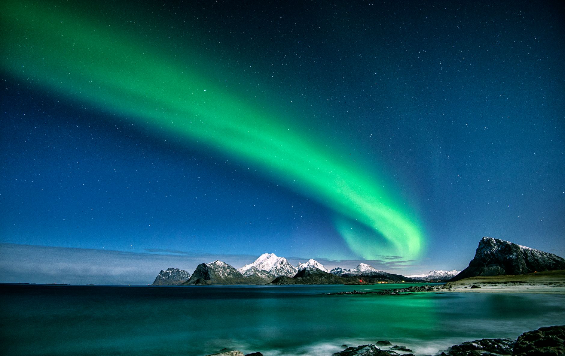 Green northern lights arcing over a calm Lofoten fjord and a dark peak reflected in still water