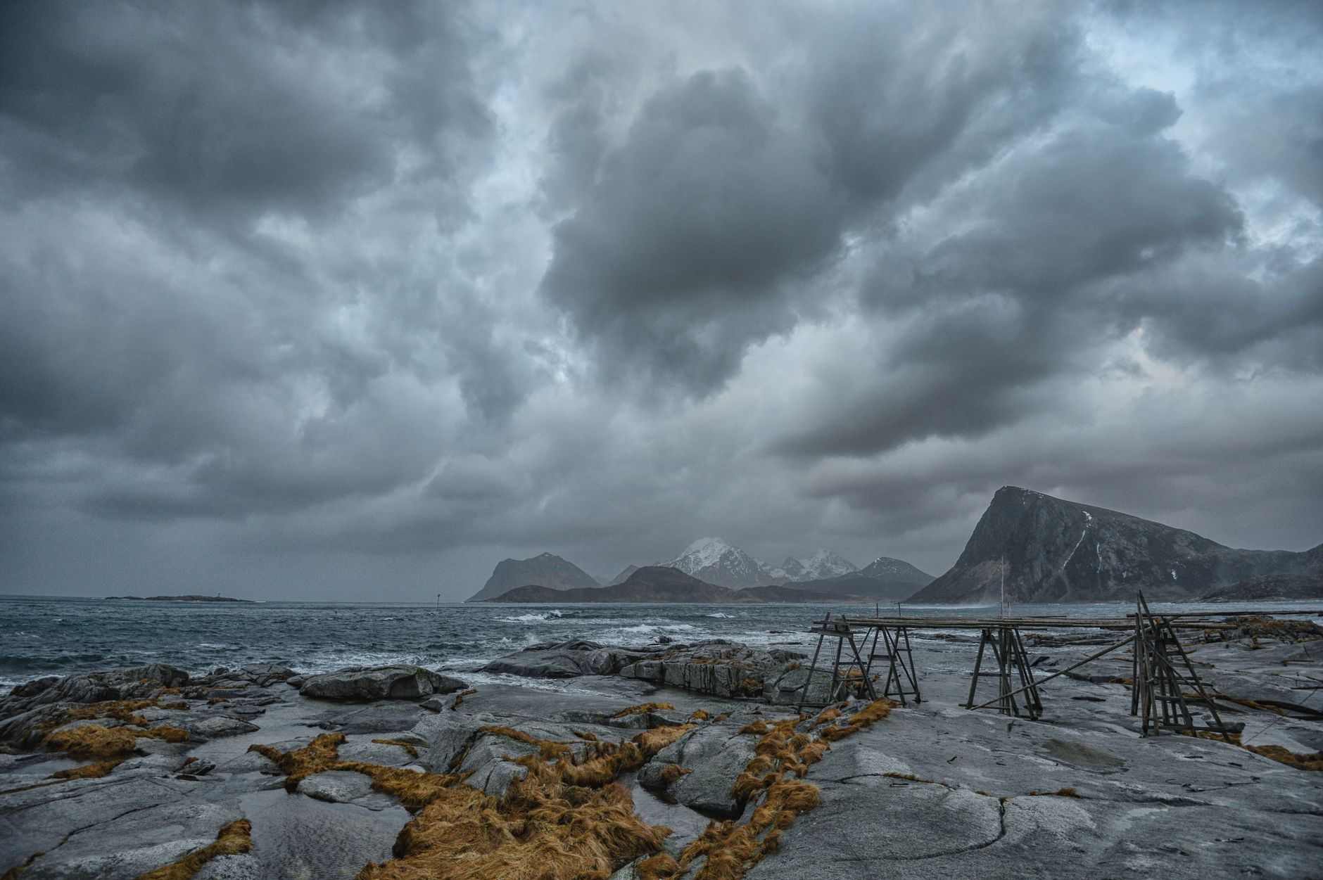 Dark cloud sitting low over grey Lofoten peaks above a cold rocky shoreline and flat sea