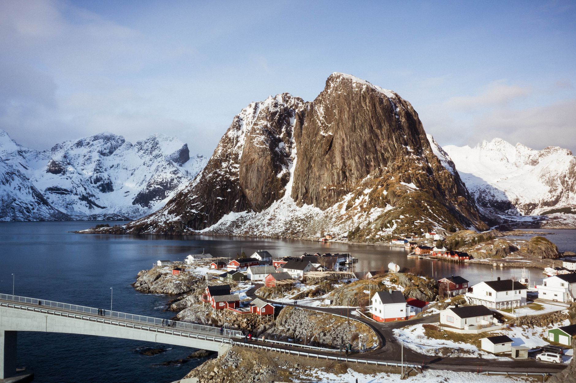 A road and bridge running past red cabins toward a sharp snow-dusted peak at Hamnoy, Lofoten, under grey sky
