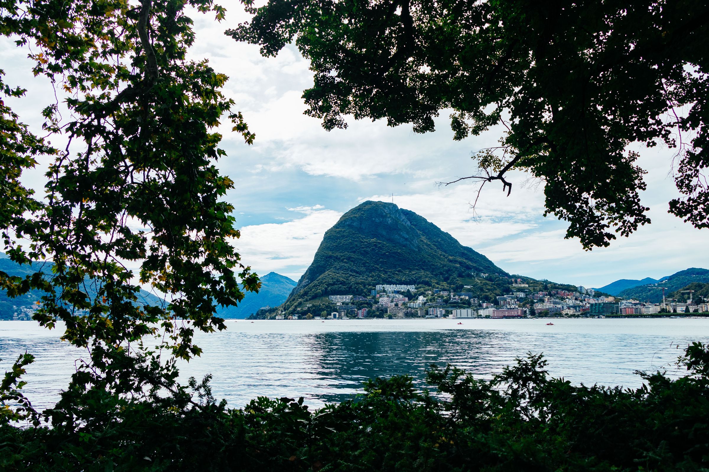 Monte San Salvatore rising across the bay of Lugano, with the houses of Paradiso along the shore below it, seen through trees on the opposite bank