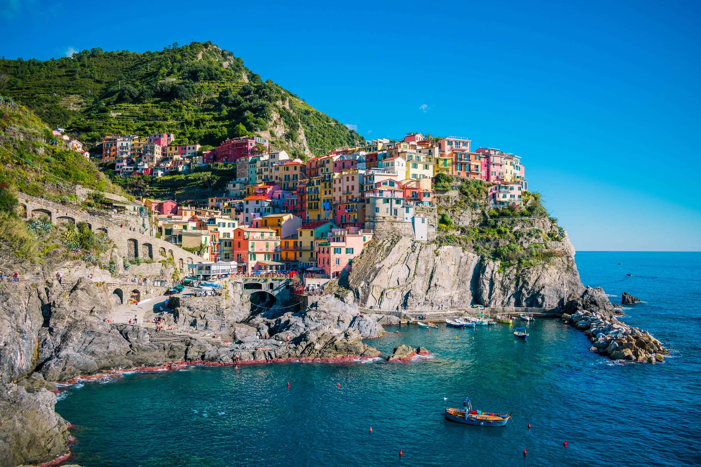 Brightly painted tall houses on a steep rock above a small rocky harbour of deep blue water, boats on the water and green terraced slopes behind under a clear blue sky