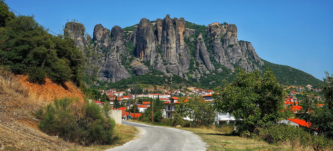A road running down to a town of red roofed houses beneath the Meteora rock wall, with a monastery on the skyline