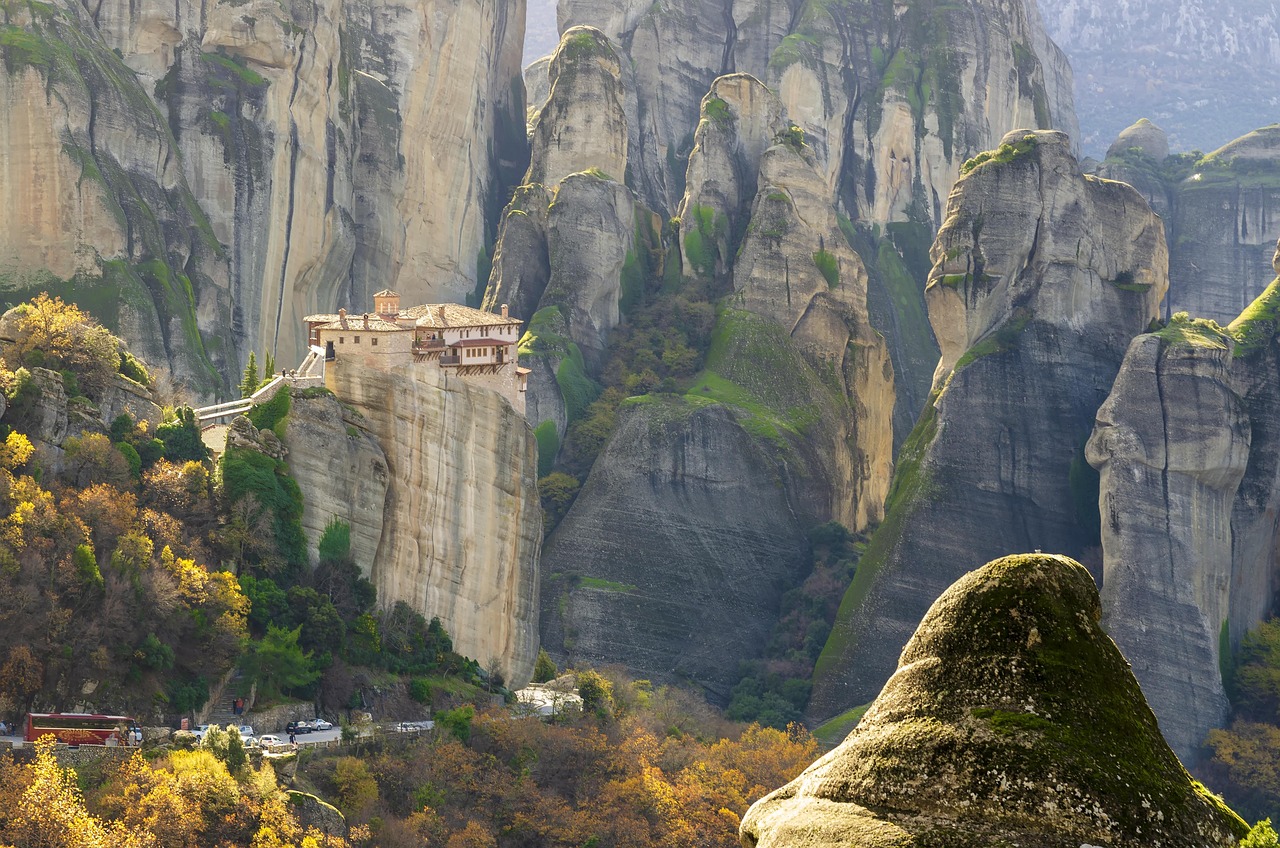 Rousanou monastery on its spur beneath the Meteora cliffs in autumn, with a road and parked coaches on the valley floor below