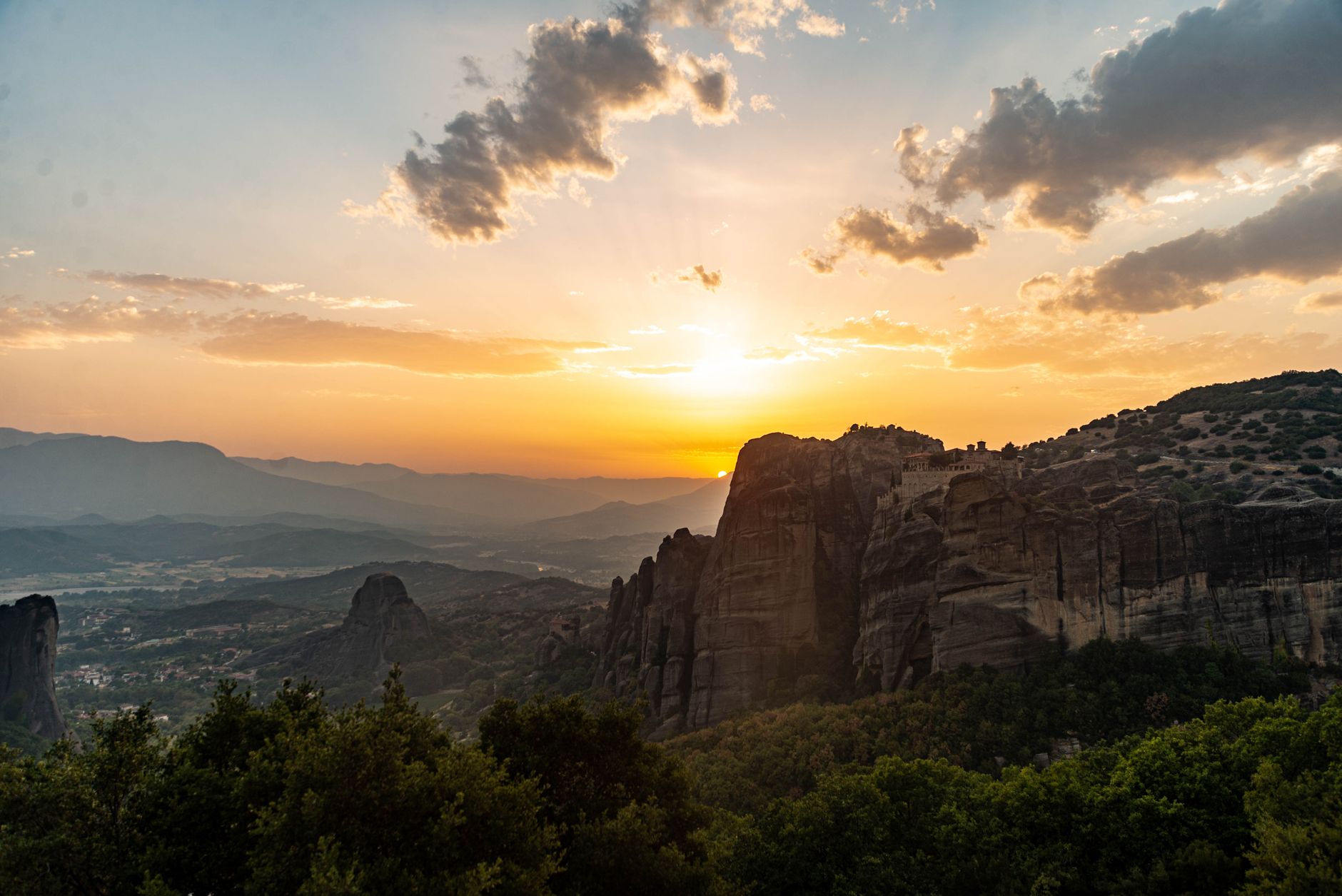 The sun setting past the Meteora rock towers with a monastery in silhouette on the ridge and the plain beyond