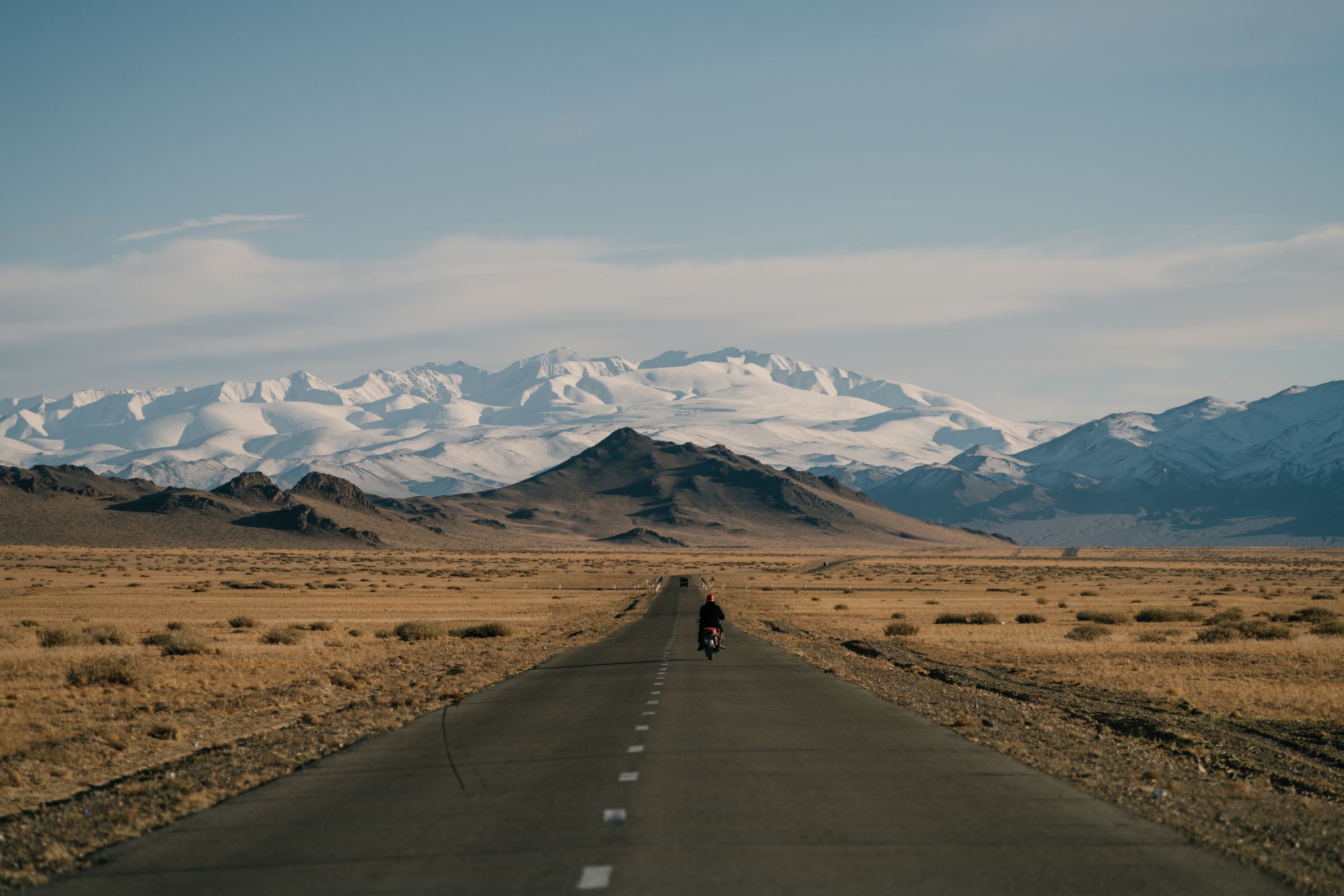A single rider on a road running toward the snow-covered peaks of the Mongolian Altai