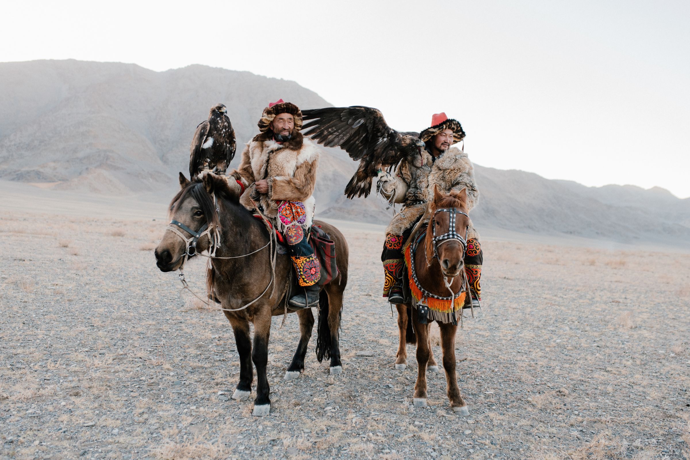 Two Kazakh eagle hunters on horseback in fur and embroidered dress, a golden eagle with wings spread, the Altai behind