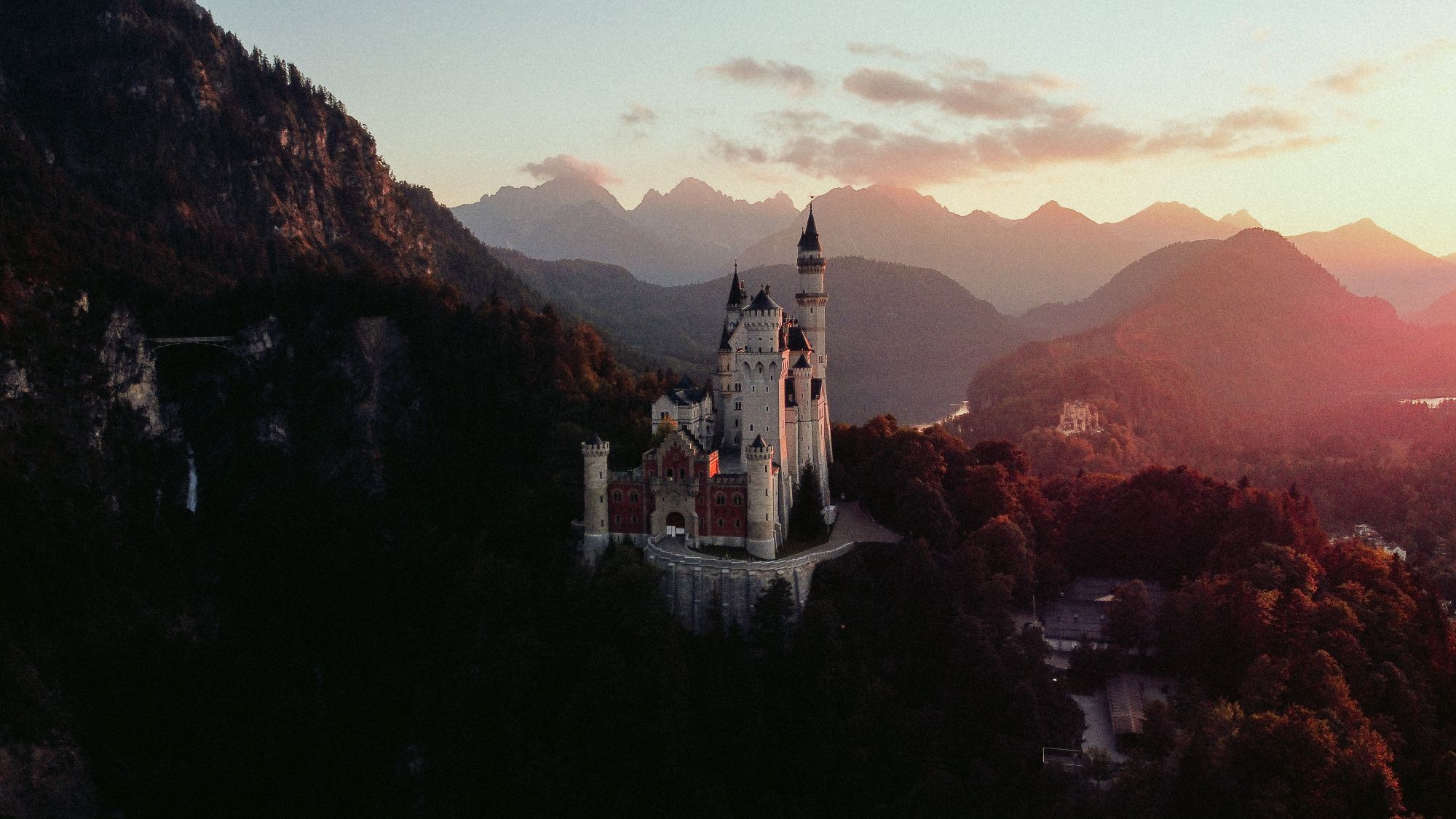Neuschwanstein in a valley at dusk, the silhouettes of the Bavarian Alps behind it in gold light