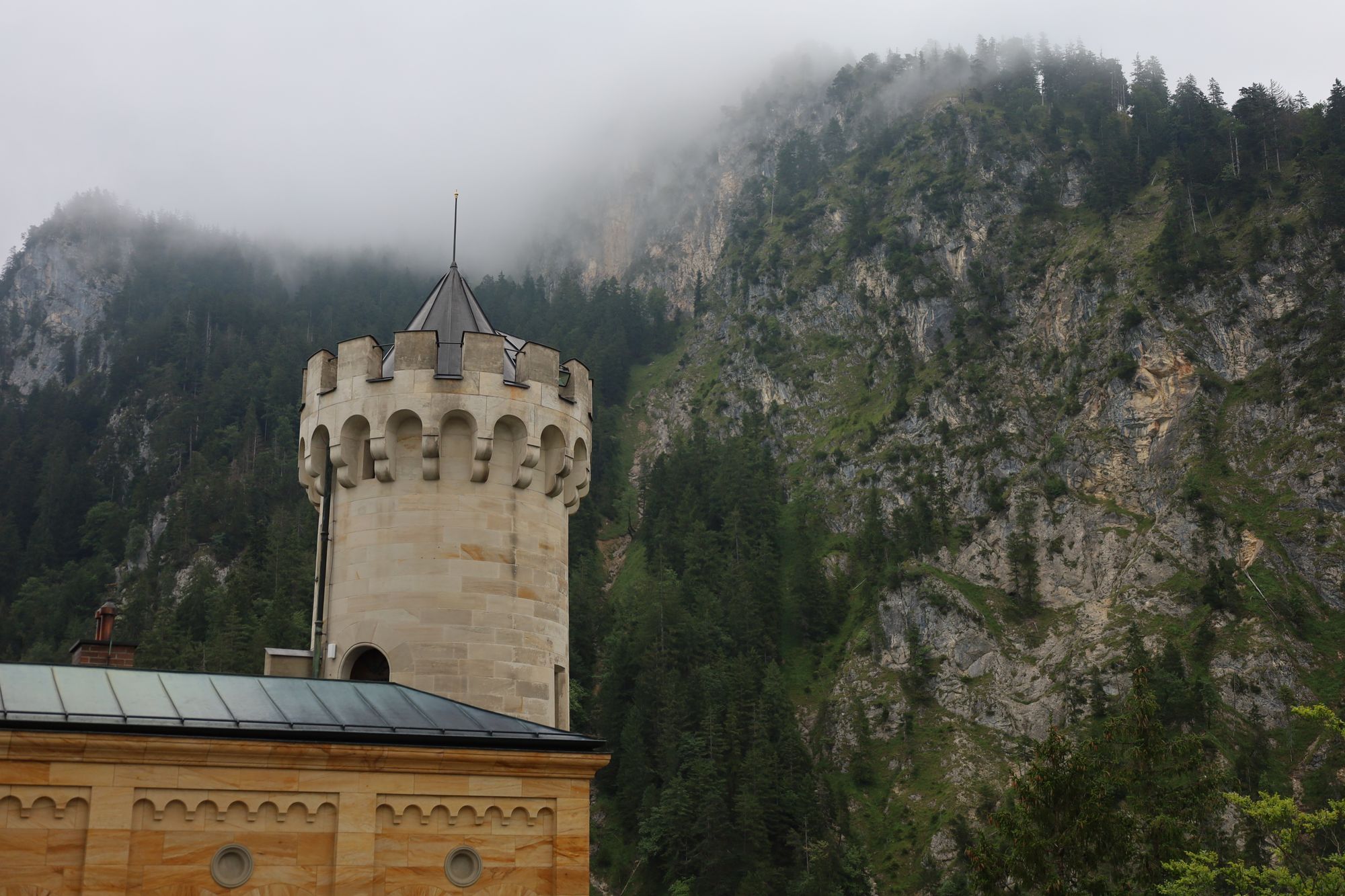 A single tower of Neuschwanstein rising in front of a mist-covered forested mountain