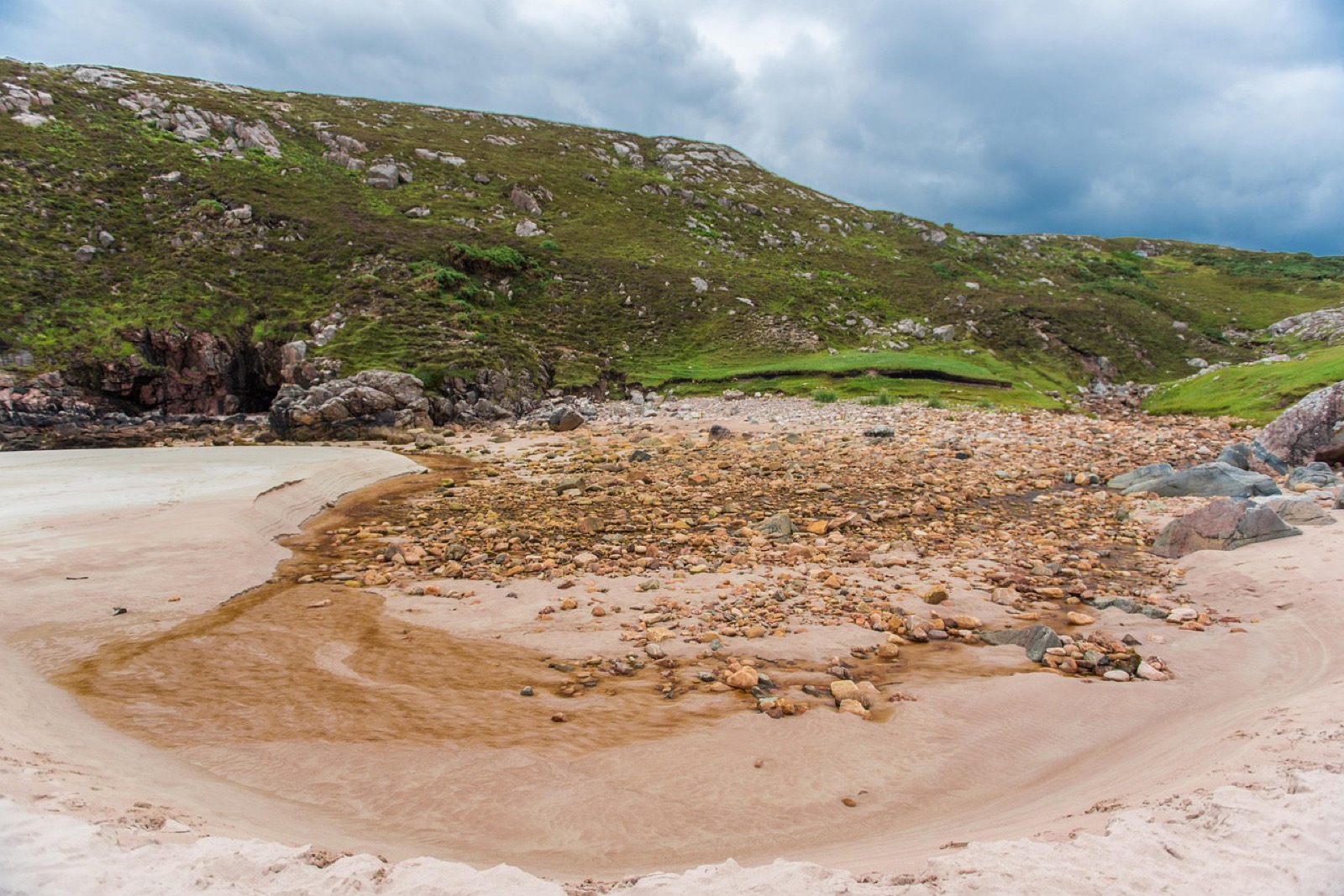 A cove of pale sand with a stream of brown peat water and rocks below a green hillside