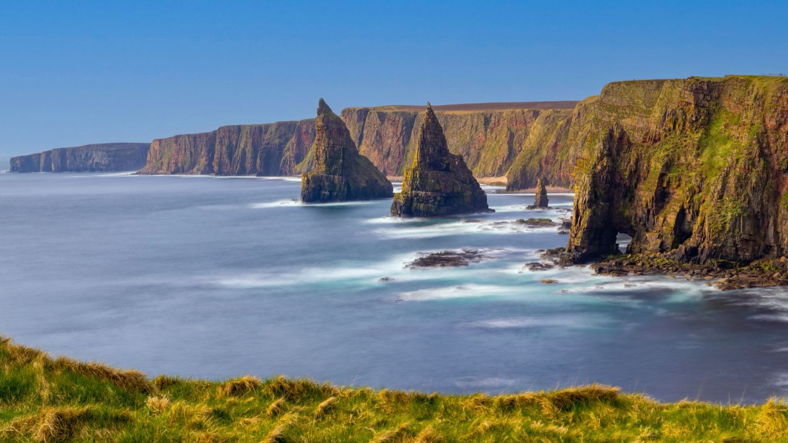 Tall sandstone sea stacks and cliffs with a rock arch, calm sea under a blue sky