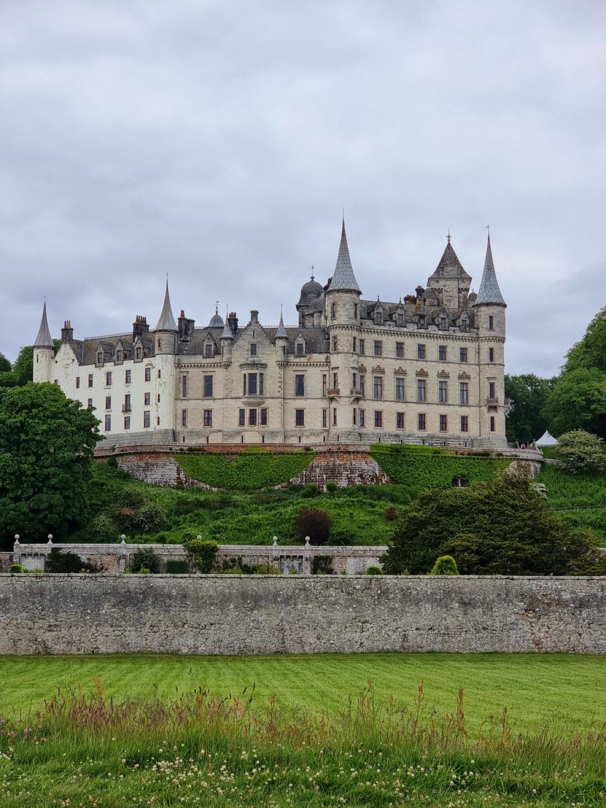 A pale turreted French-style castle above terraced gardens behind a stone wall, grey sky