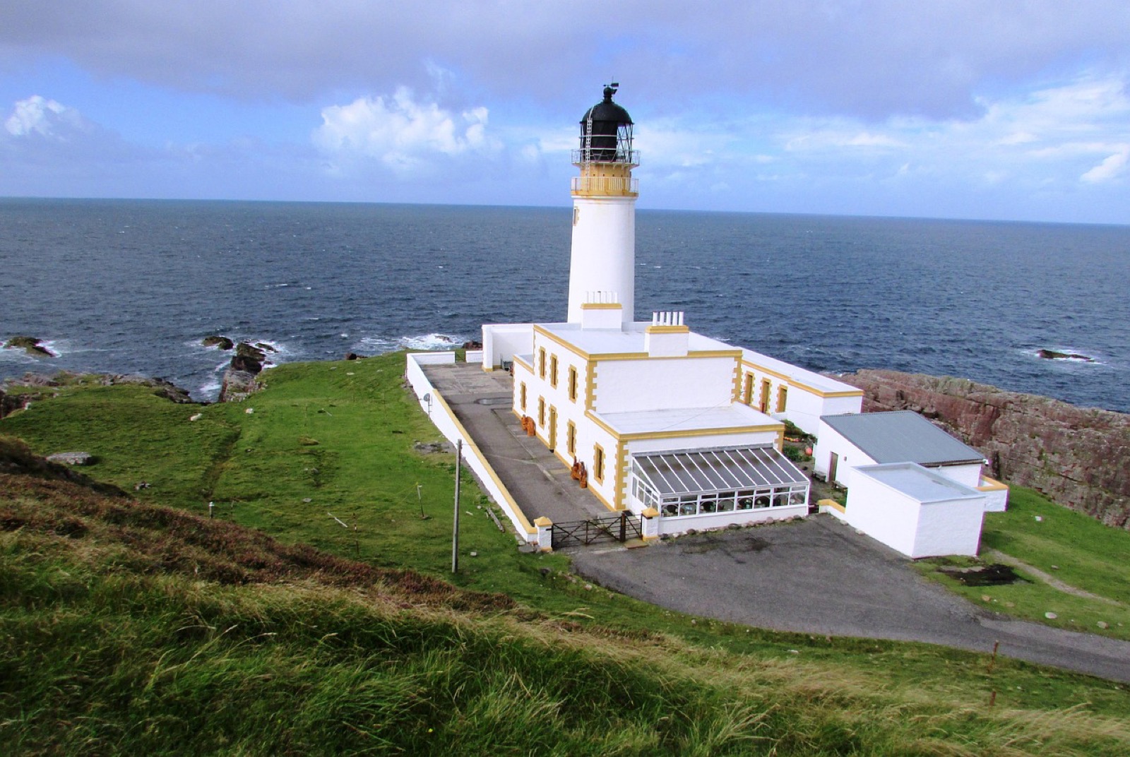 A white lighthouse with yellow trim and keepers' buildings on a green headland above a dark sea