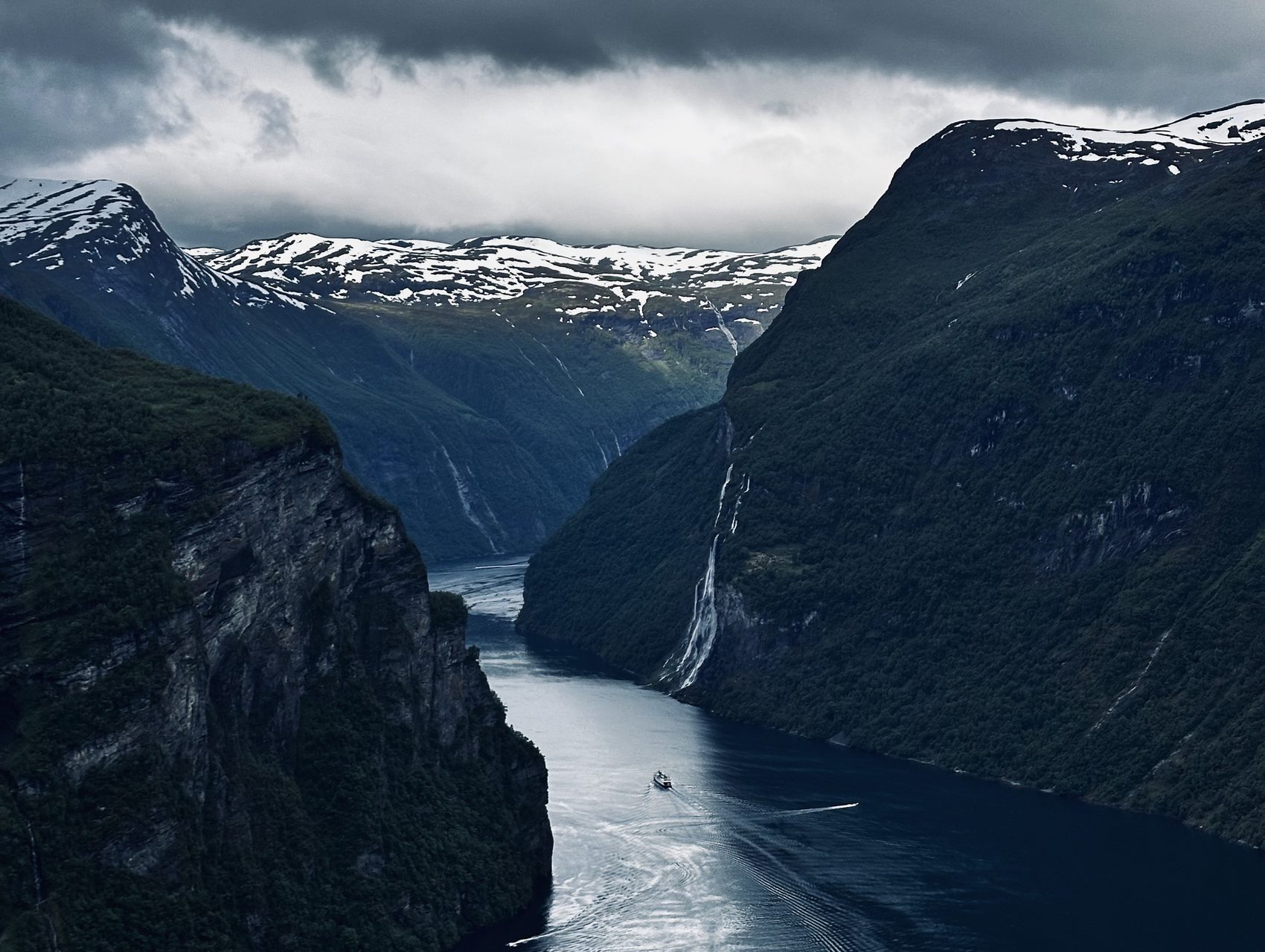 A narrow fjord between sheer cliffs, a small boat threading through beneath snow-capped peaks and waterfalls