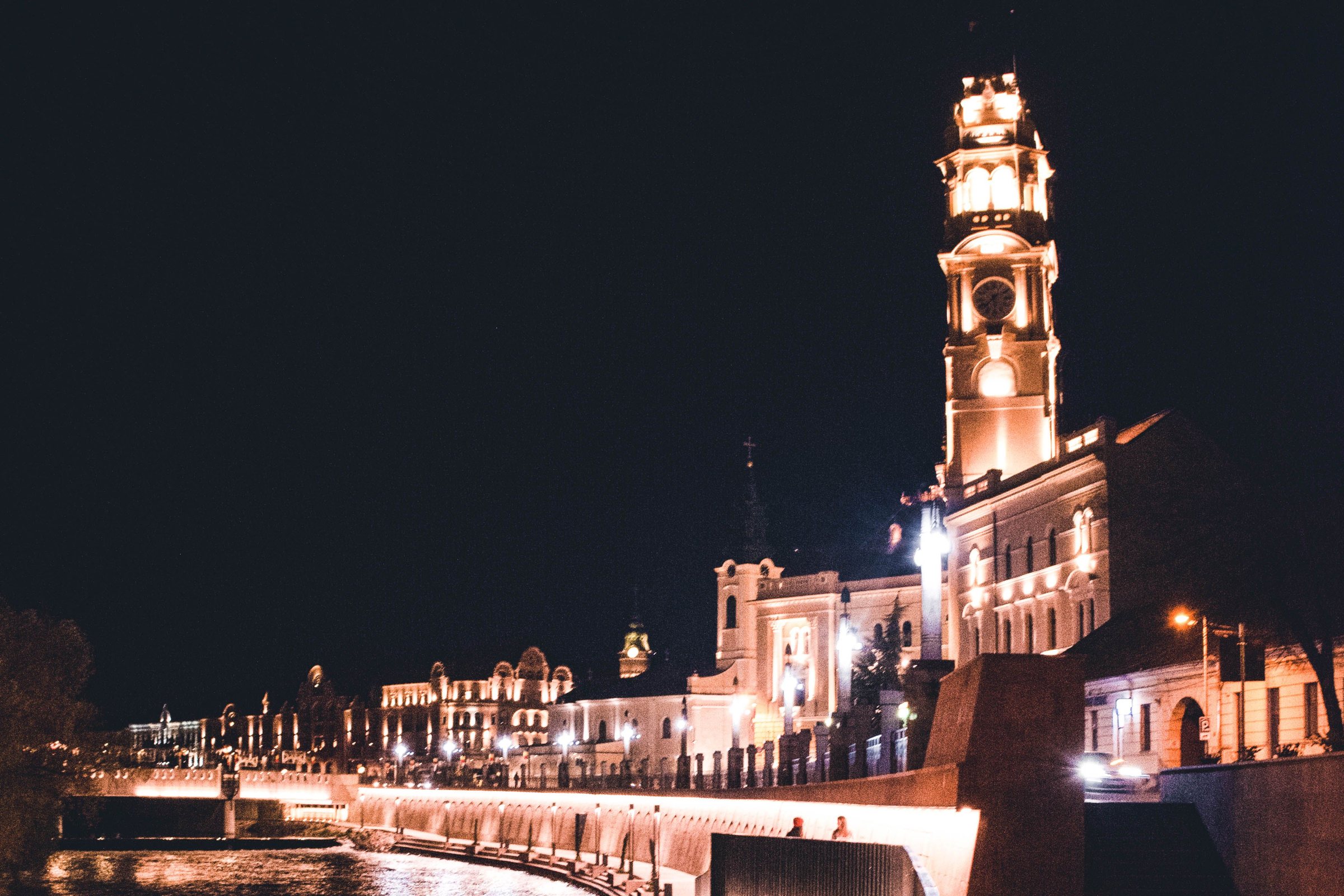 Oradea's city hall clock tower lit at night above the curving embankment of the Crișul Repede, with the river in the foreground