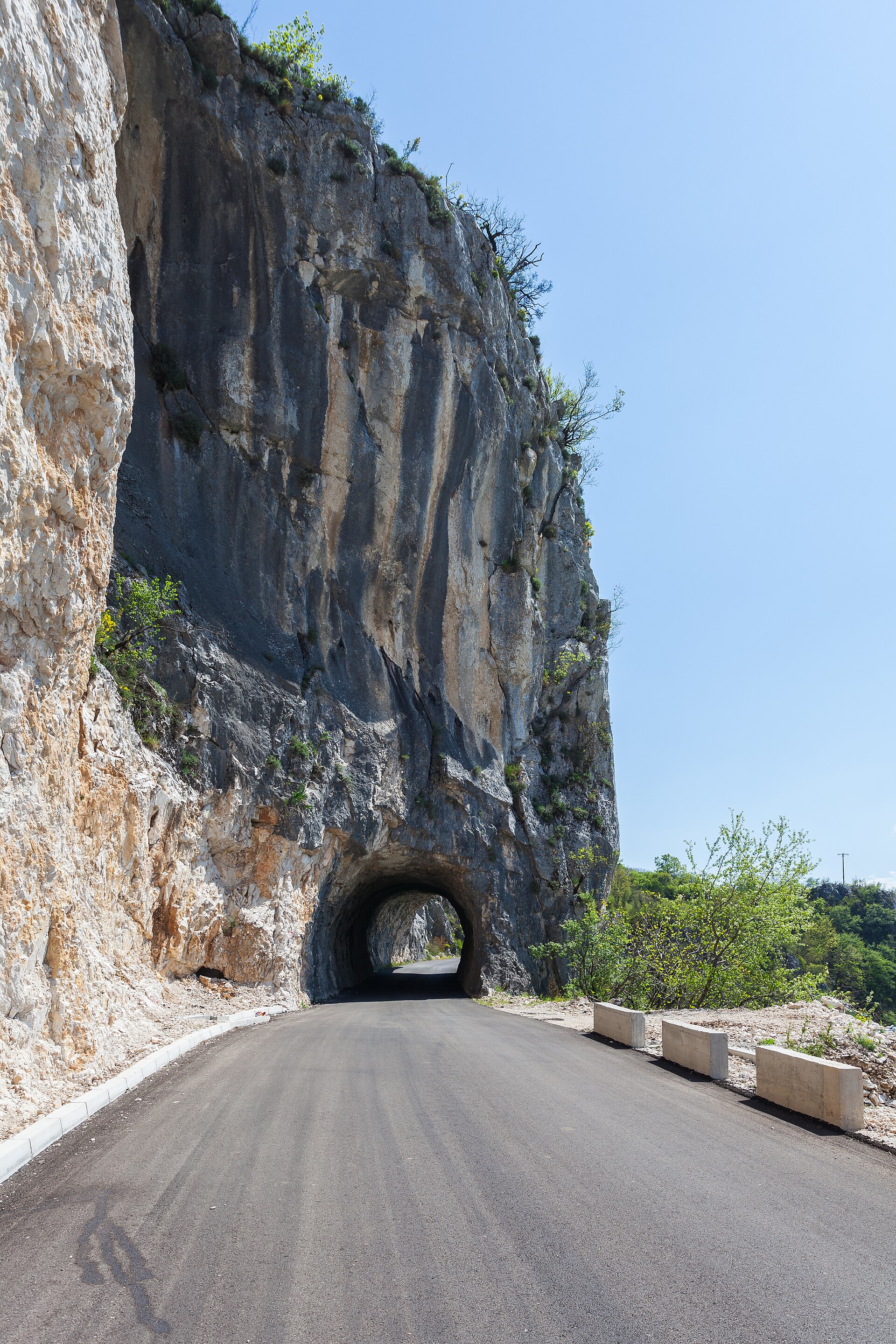 A narrow paved road cut along a cliff face at Ostrog, passing through a short rock tunnel with a drop to the valley