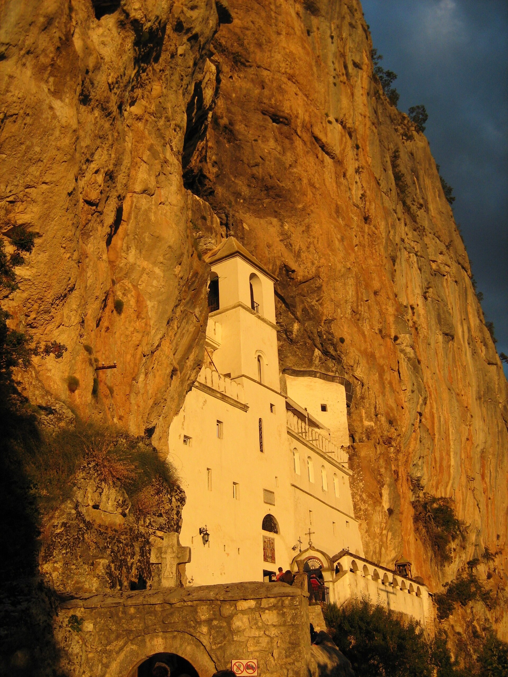 The white upper monastery of Ostrog lit warm against the orange rock hollow it is built into, the cliff rising dark above it