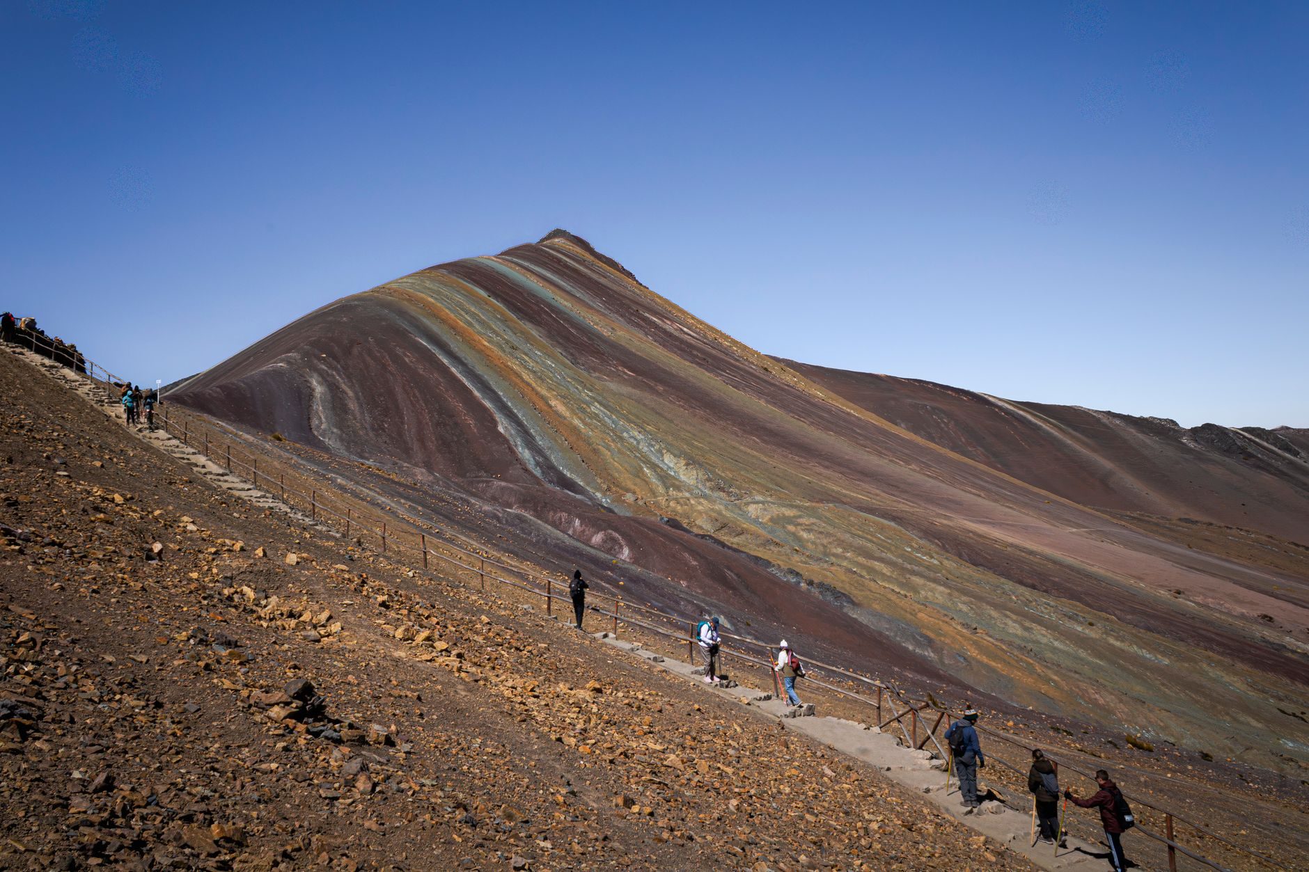 A line of walkers on the red and ochre track climbing towards the striped ridge of Vinicunca under a blue sky