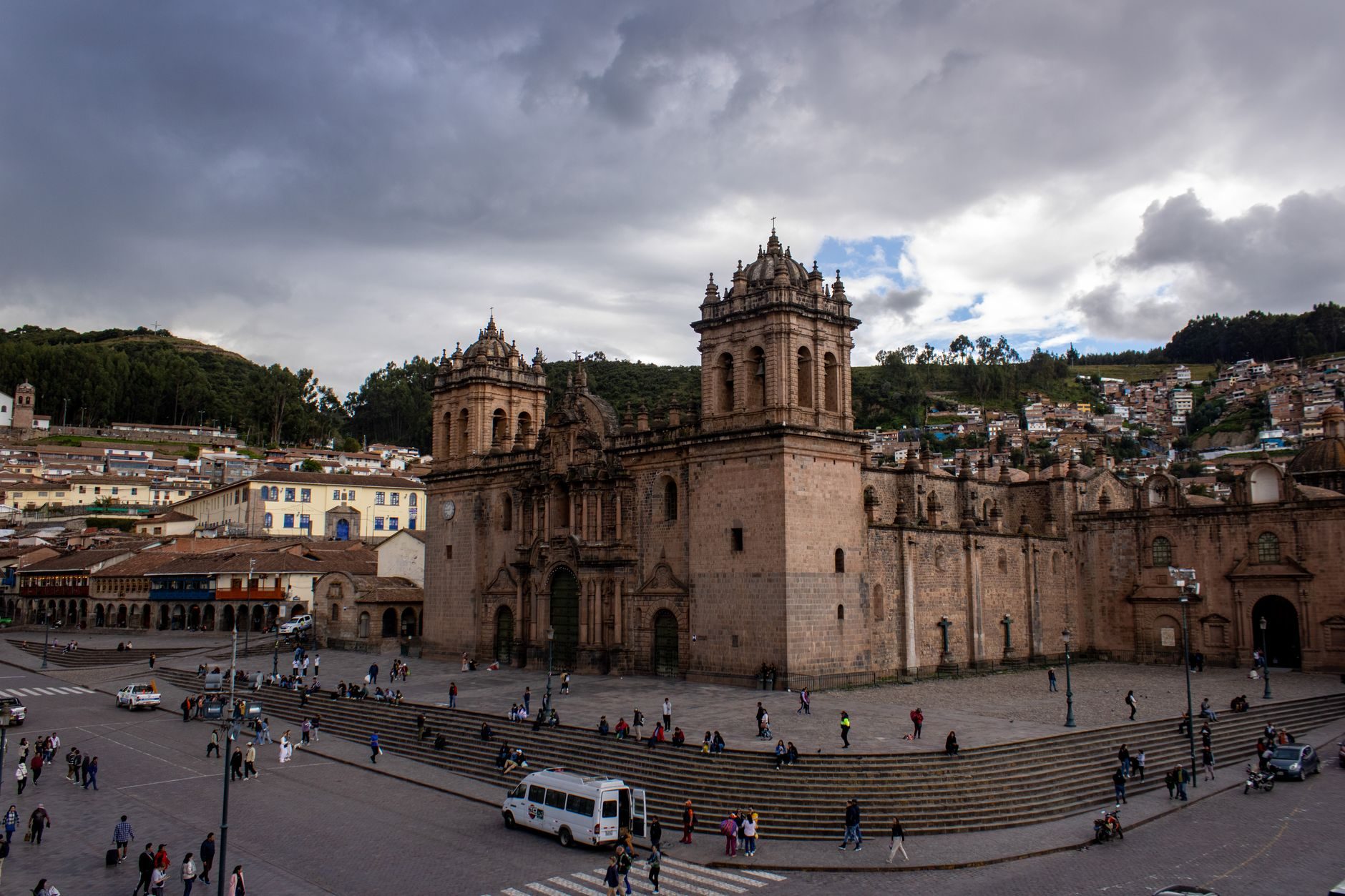 The cathedral of Cusco across the Plaza de Armas under a heavy sky, its two towers and the arcaded square in front with people crossing it