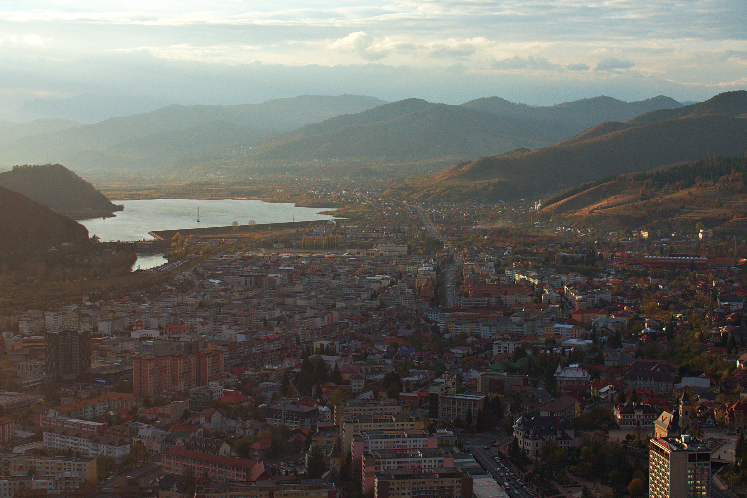 Piatra Neamț from the hills above it, apartment blocks and red roofs filling the valley floor, Bâtca Doamnei lake and its dam on the left and wooded ridges behind