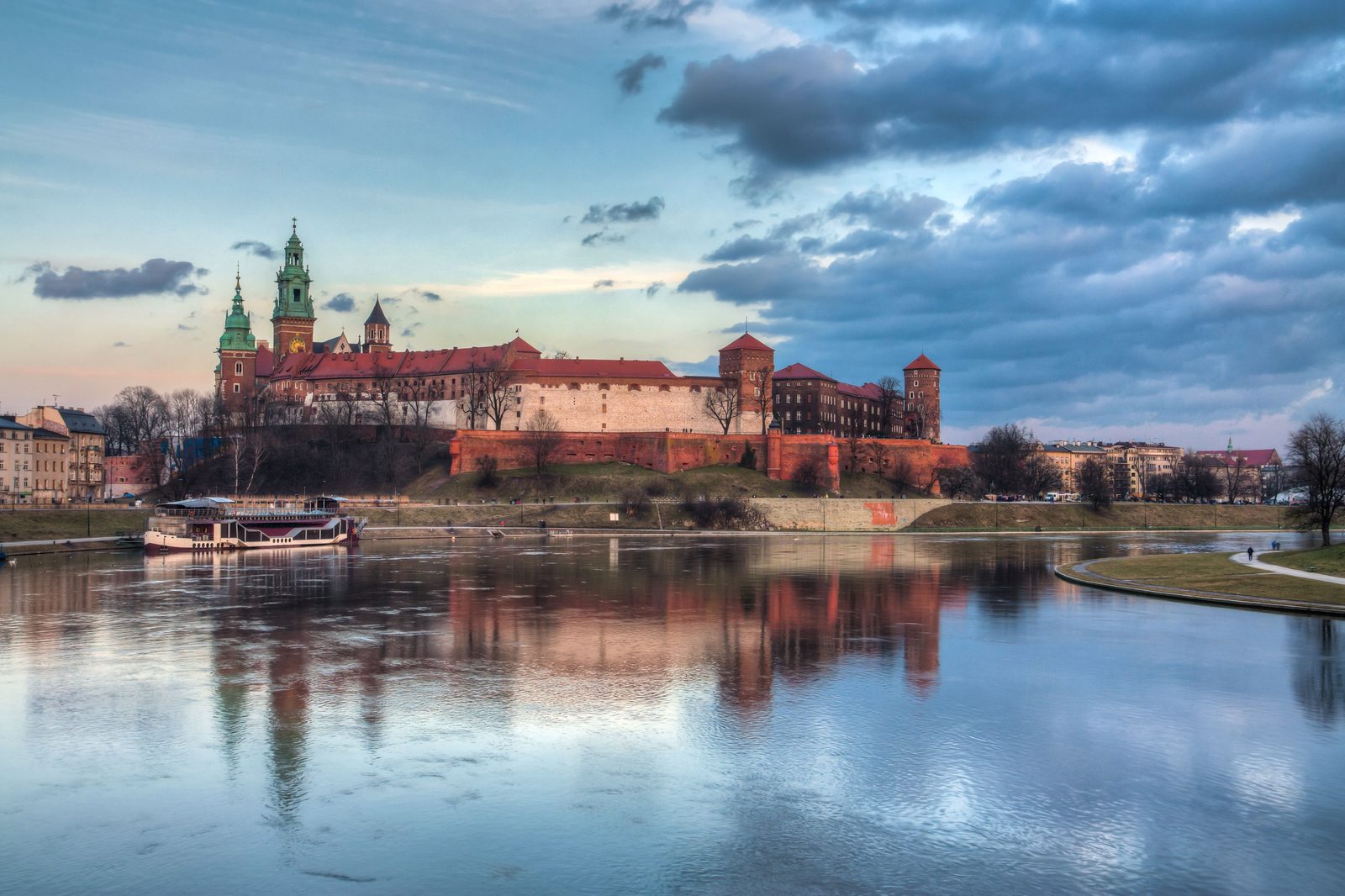 Wawel Castle and its cathedral towers on the hill above the Vistula in Kraków, reflected in the river at sunset under a cloudy sky