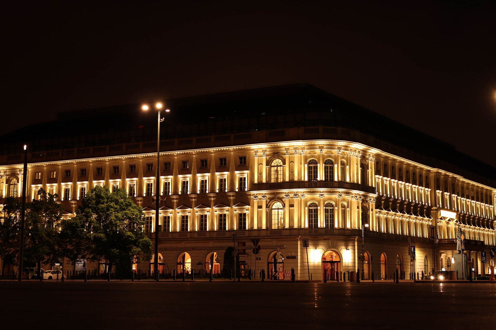 The Raffles Europejski in Warsaw at night, its long neoclassical front lit gold above an empty, rain-wet street