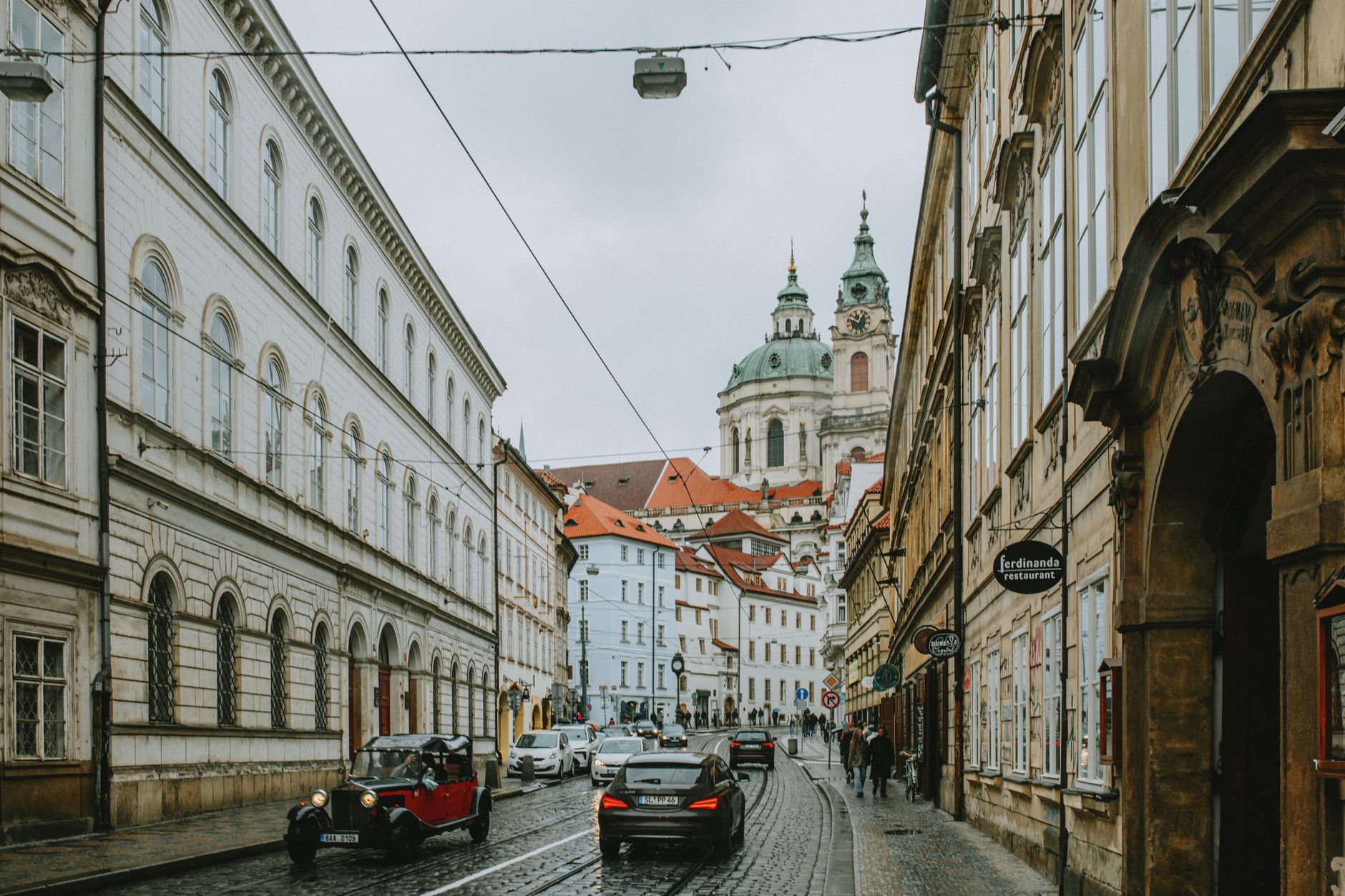 A wet Malá Strana street with tram tracks and cars, the green dome of St Nicholas behind