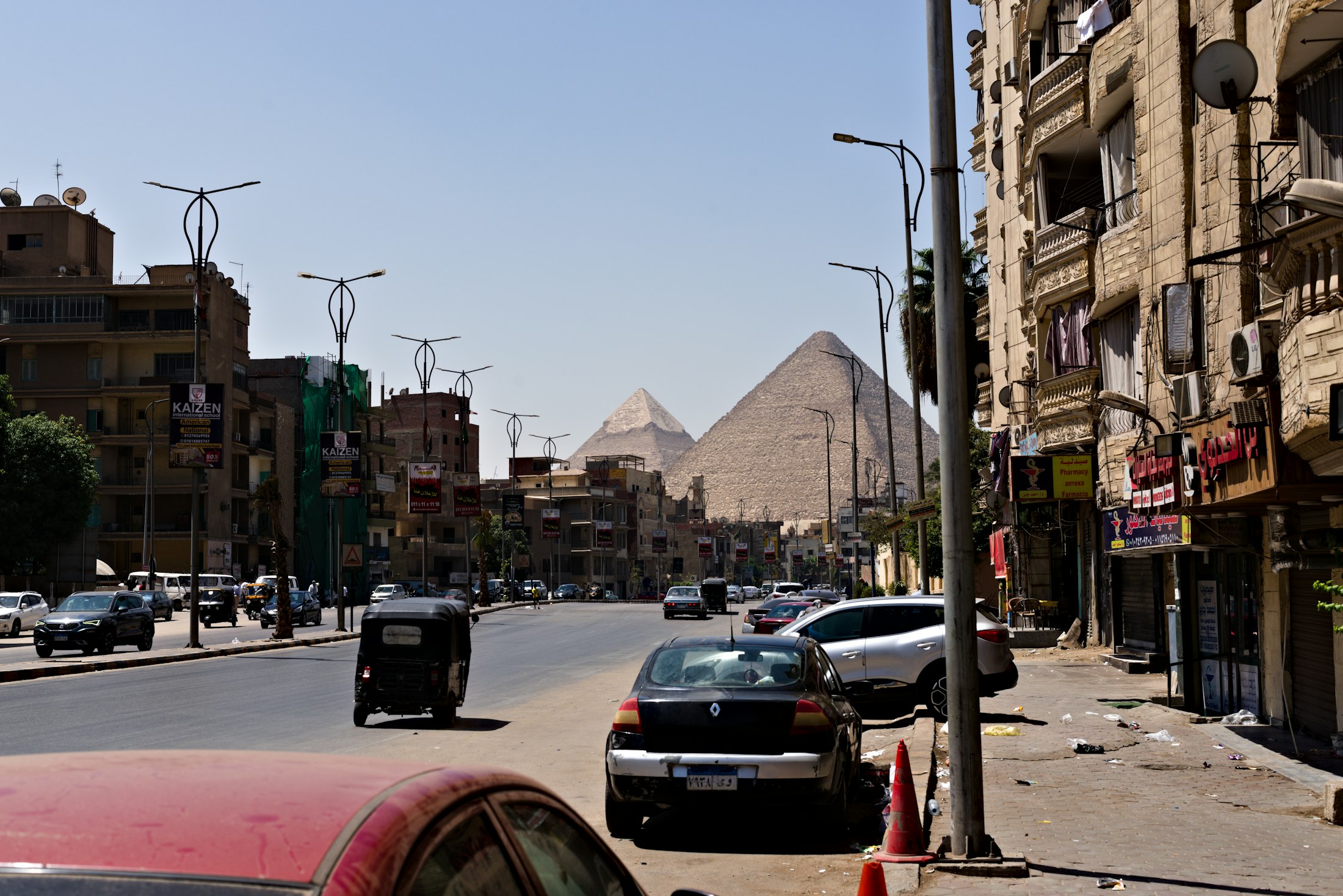An ordinary Cairo street with cars and shops, the Pyramids of Giza visible over the rooftops at the end of it