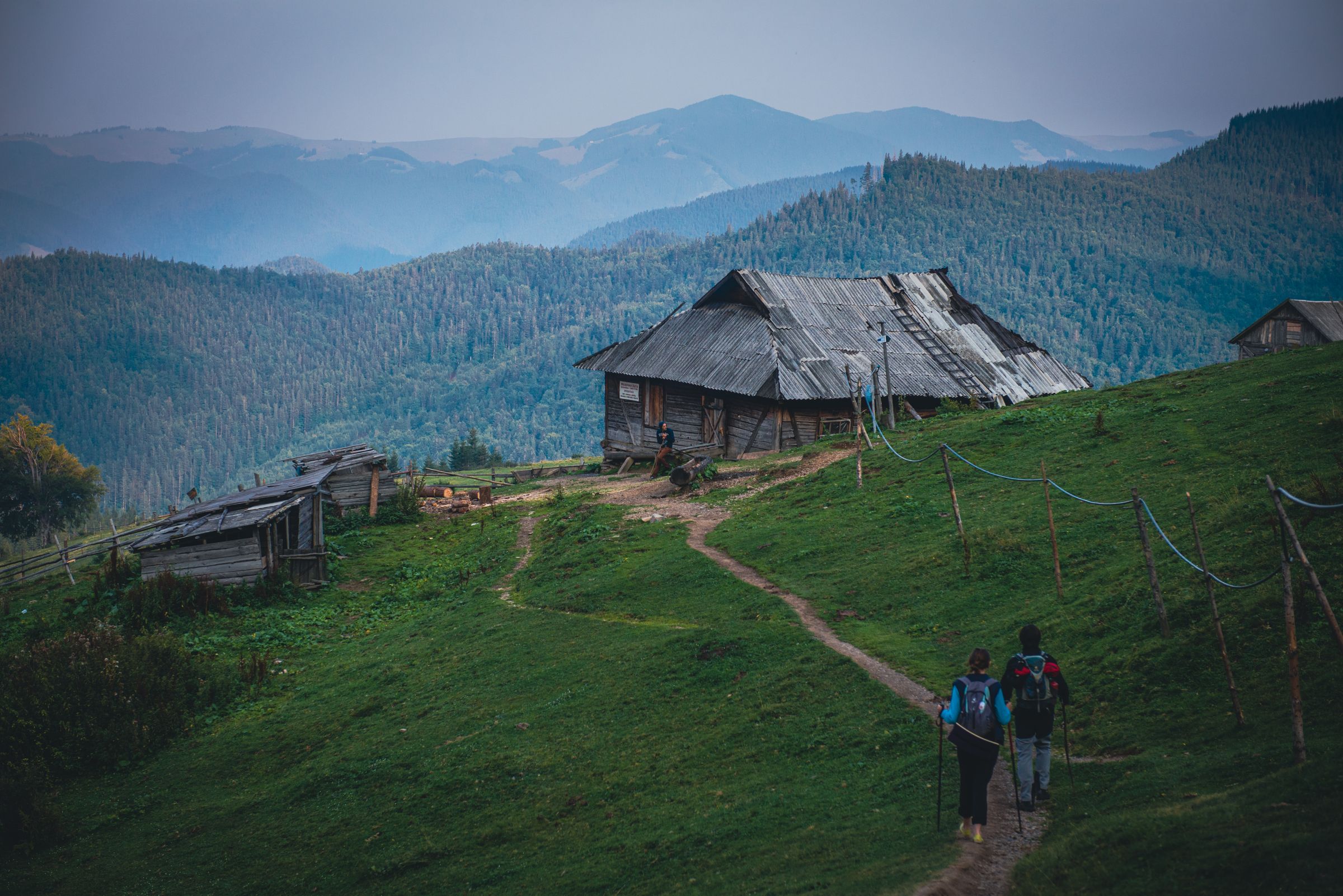 Two walkers with packs climbing a grass path to a shepherd’s hut, forested Carpathian ridges behind