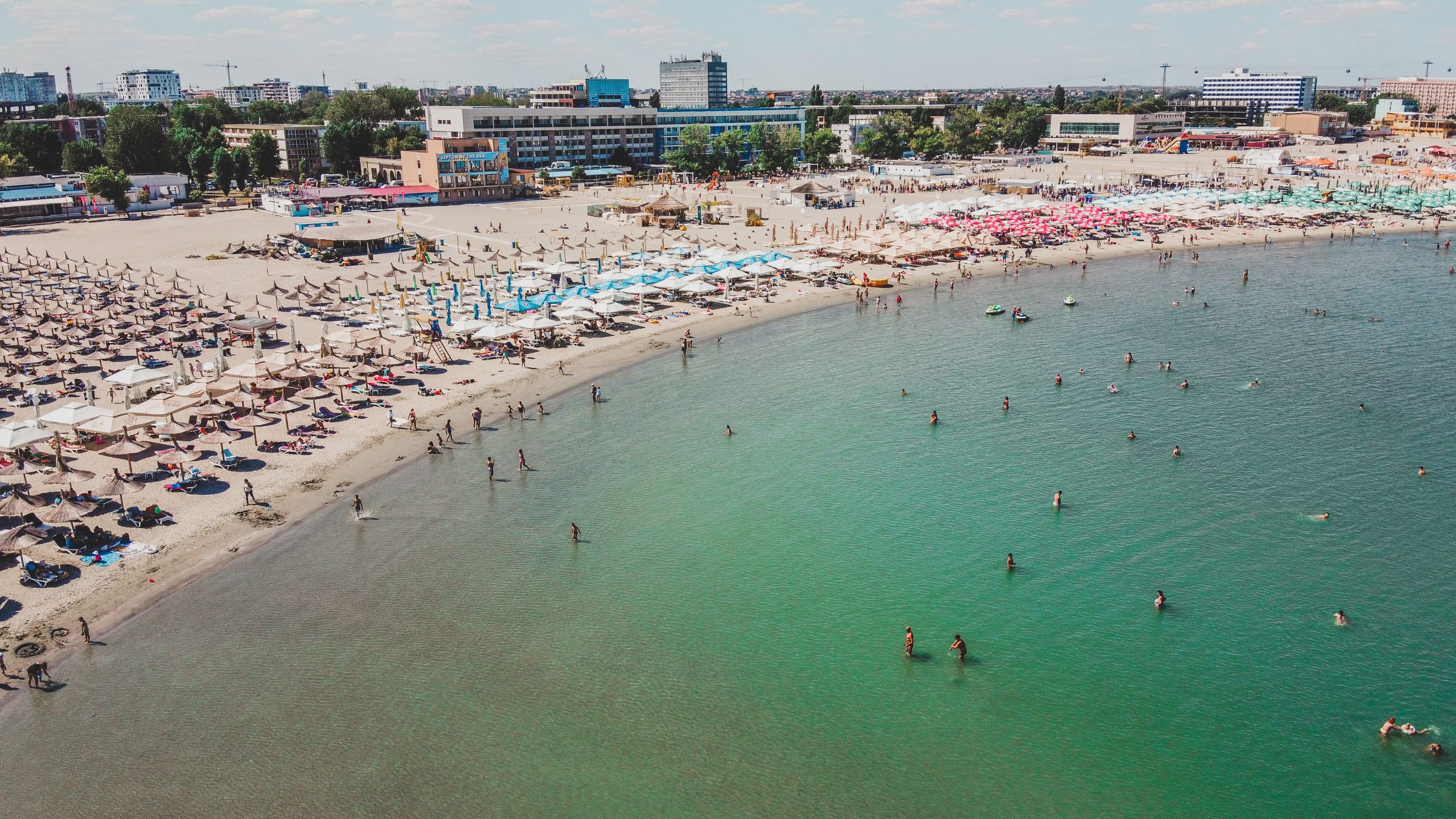 An aerial view of a busy Constanța beach in summer, thatched umbrellas on the sand and swimmers in shallow green water
