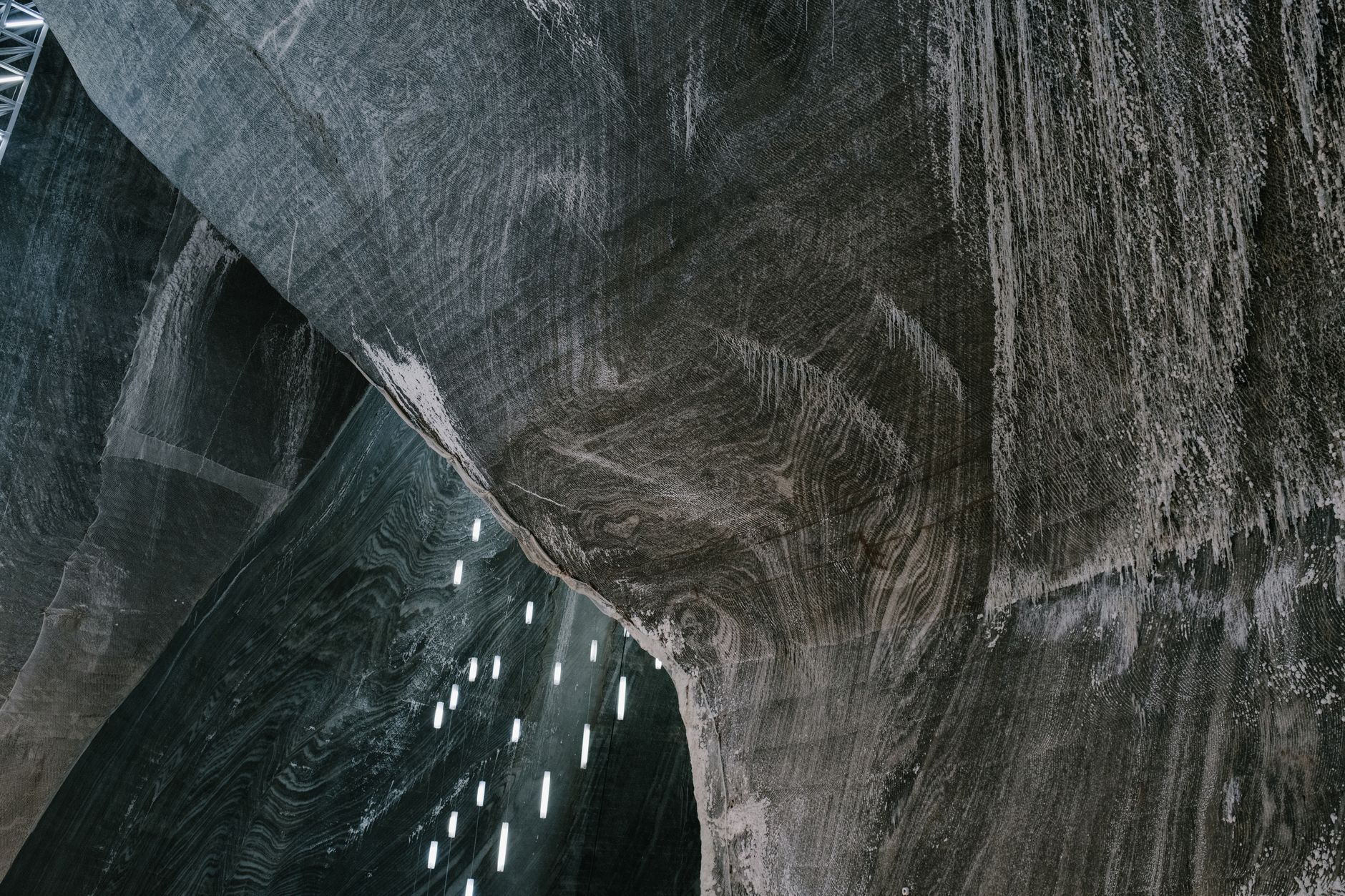 A vast curved wall of grey and brown banded salt, with small light tubes hanging in the distance