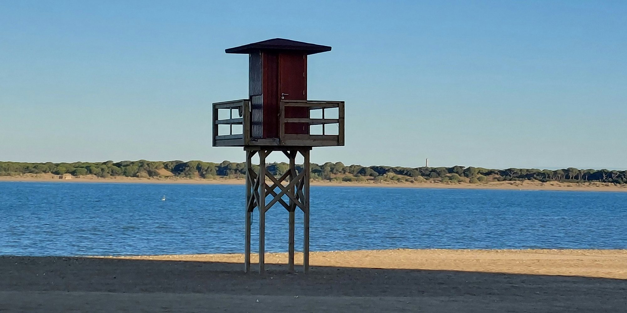 A wooden lifeguard tower on the beach at Sanlúcar de Barrameda, with the Guadalquivir behind it and the pine woods and dunes of Doñana on the far bank