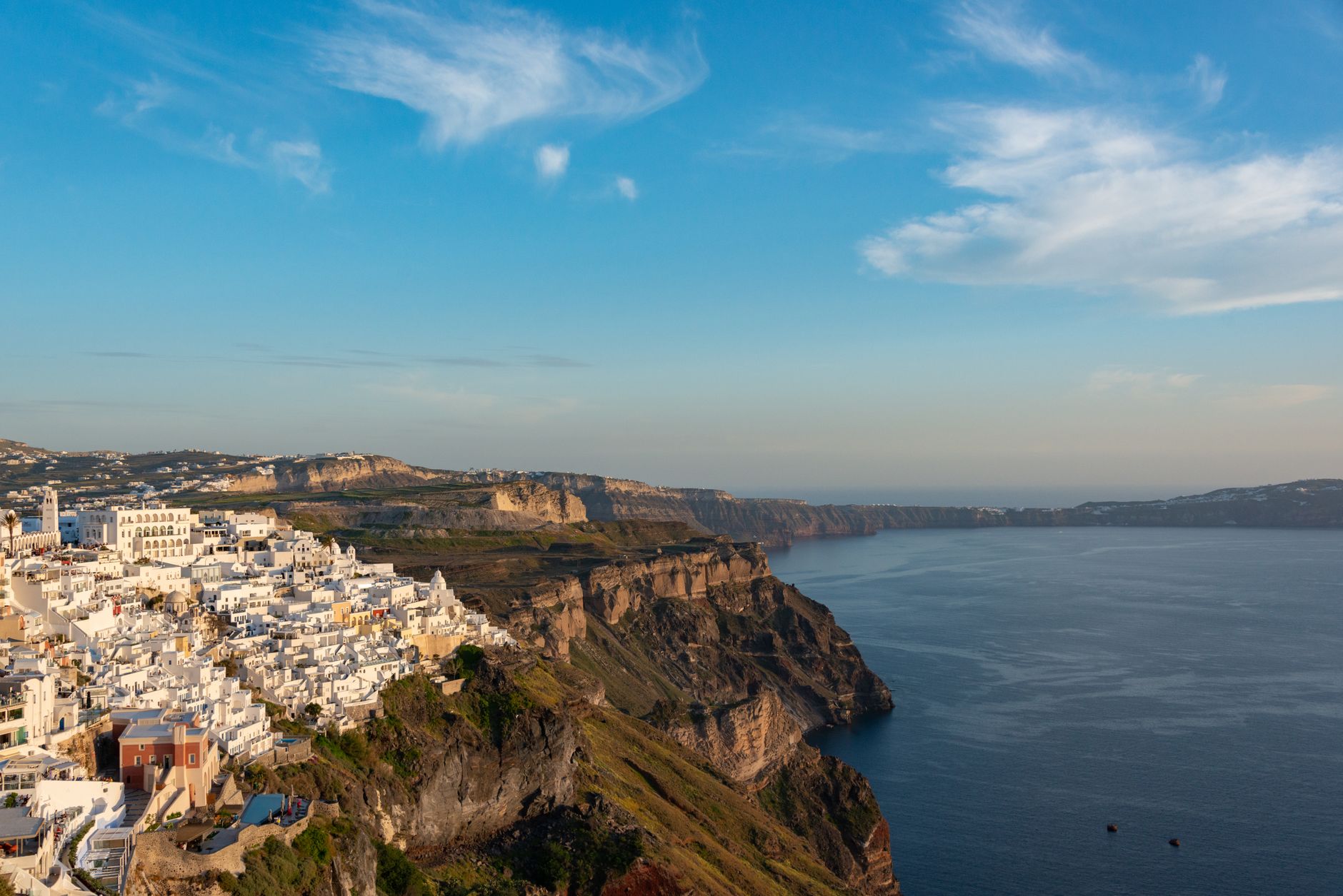 The caldera cliffs of Santorini falling to a deep blue sea, white buildings along the rim and a dark volcanic islet in the distance