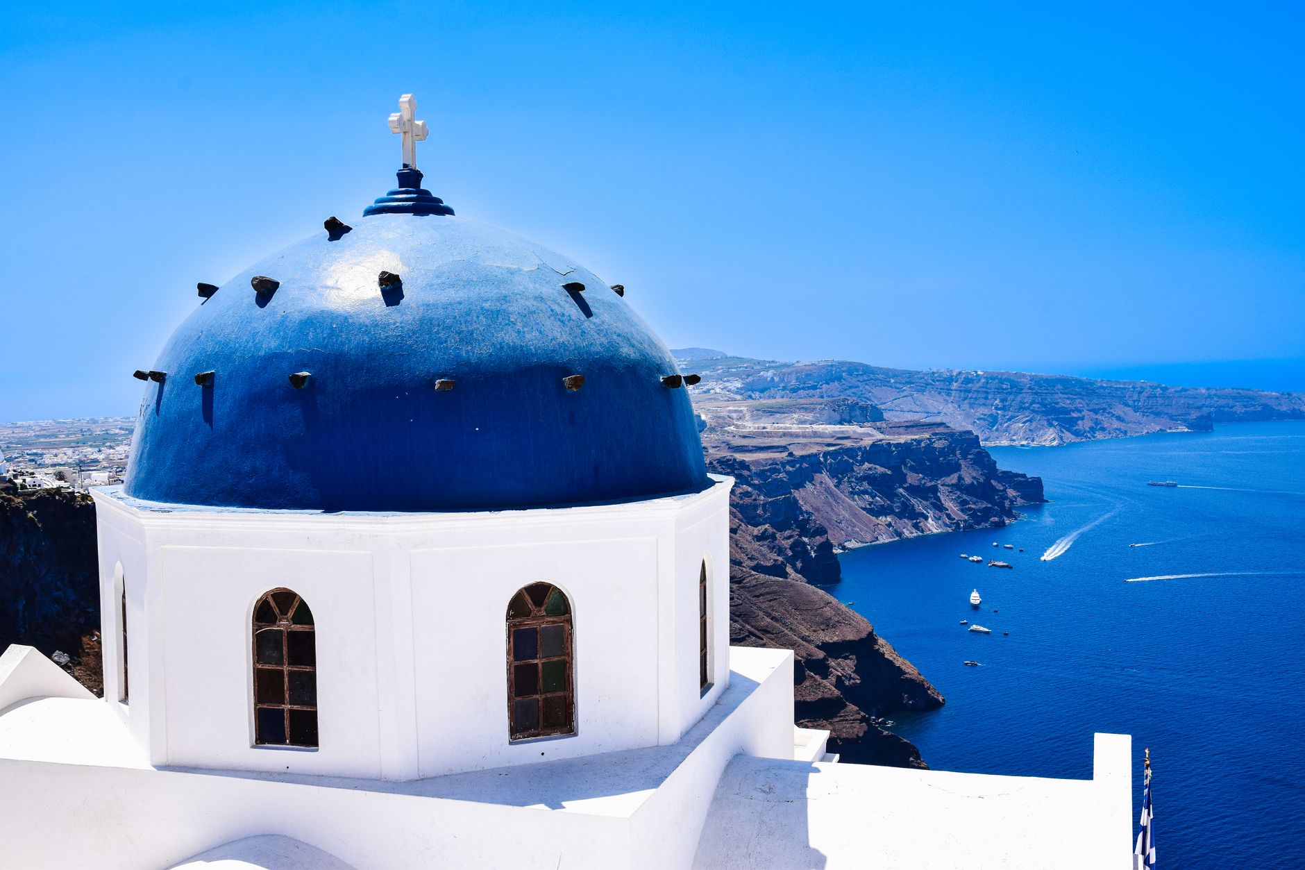A blue-domed white church on the Santorini cliff edge above the caldera, boats on the sea far below