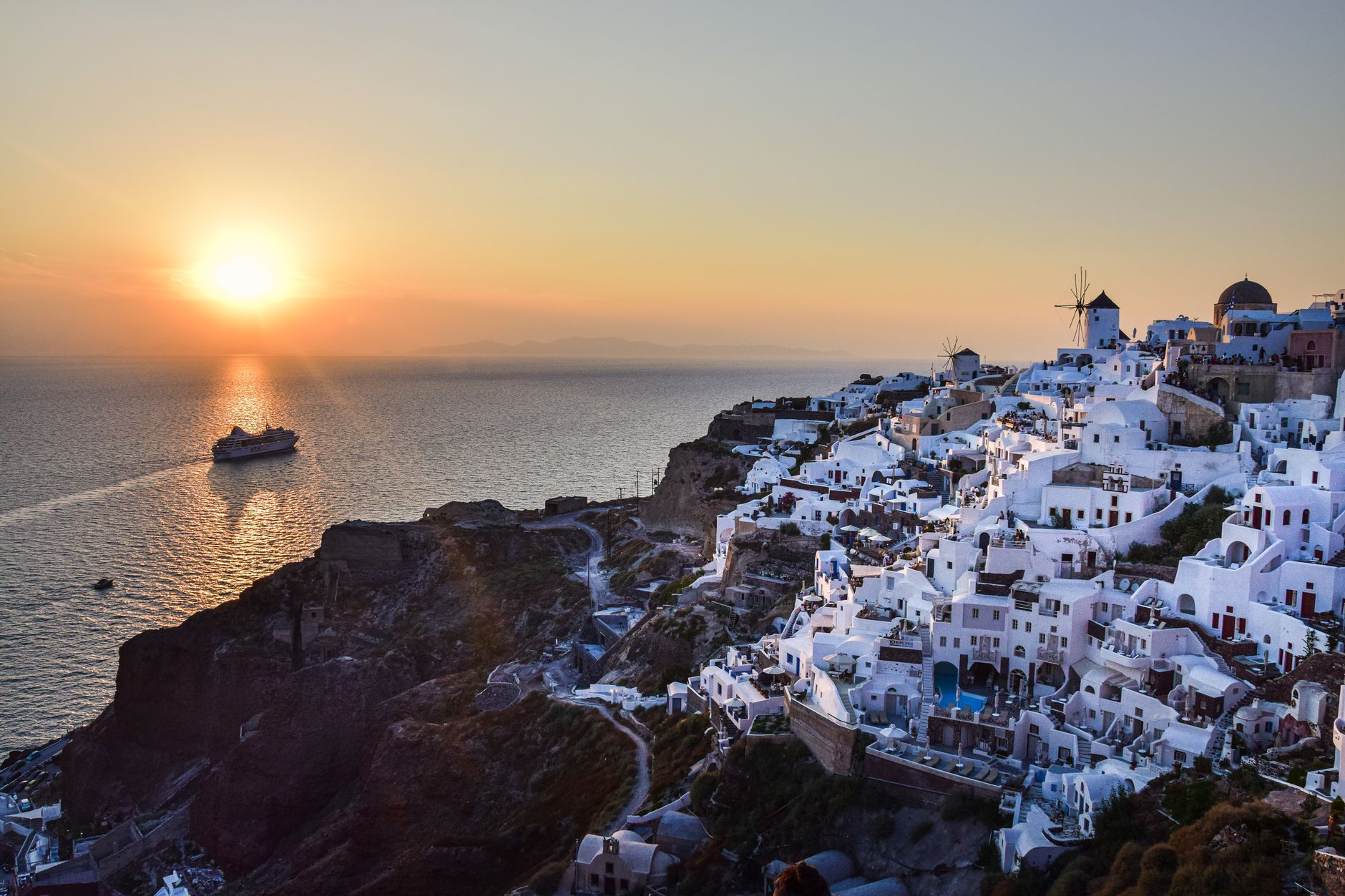 The sun setting over the sea beyond Oia, the white town on the Santorini cliff turning gold, a boat on the water