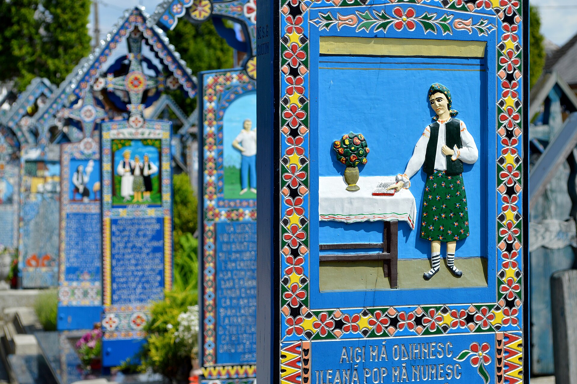 A carved and painted cross panel showing a woman standing at a cloth-covered table with a flowering plant and an open book