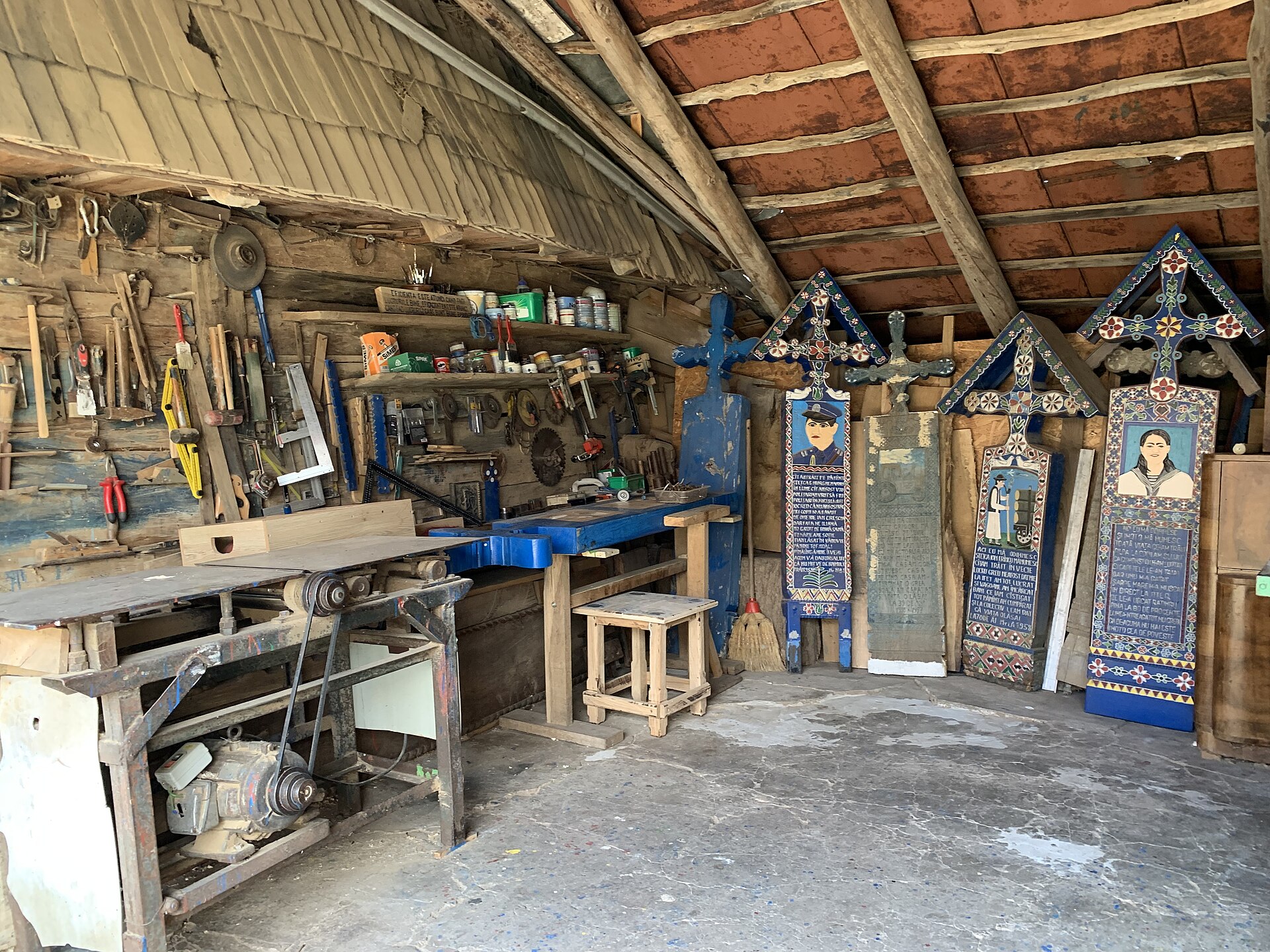 A timber workshop with hand tools on the wall and cross-shaped boards leaning against the planks, three painted and one bare