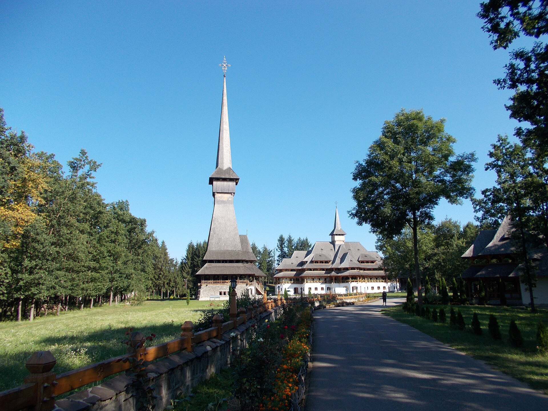 A very tall shingled wooden church spire above a monastery complex at the end of a tree-lined avenue