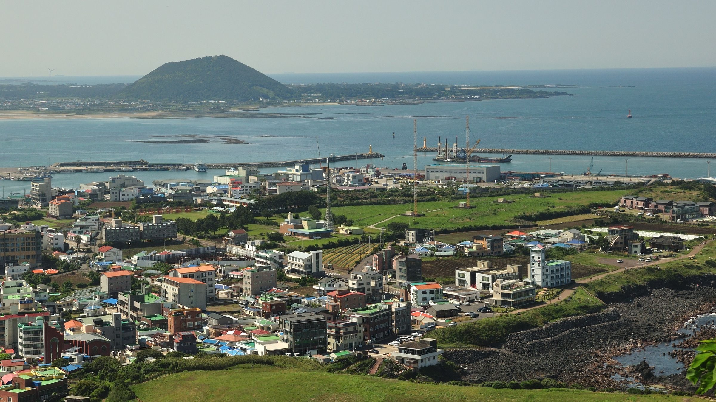 Aerial view of a harbour town and dark lava coast on Jeju's south-east, with a conical hill and a low island across the water