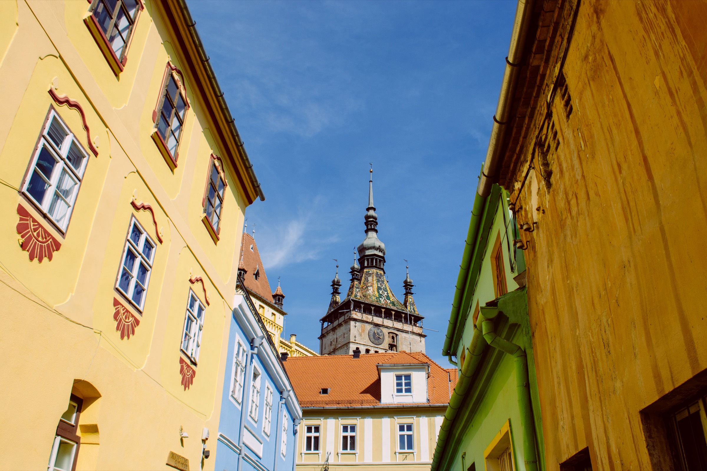 The Clock Tower of Sighișoara seen up a narrow lane of yellow, blue and green citadel houses under a clear sky
