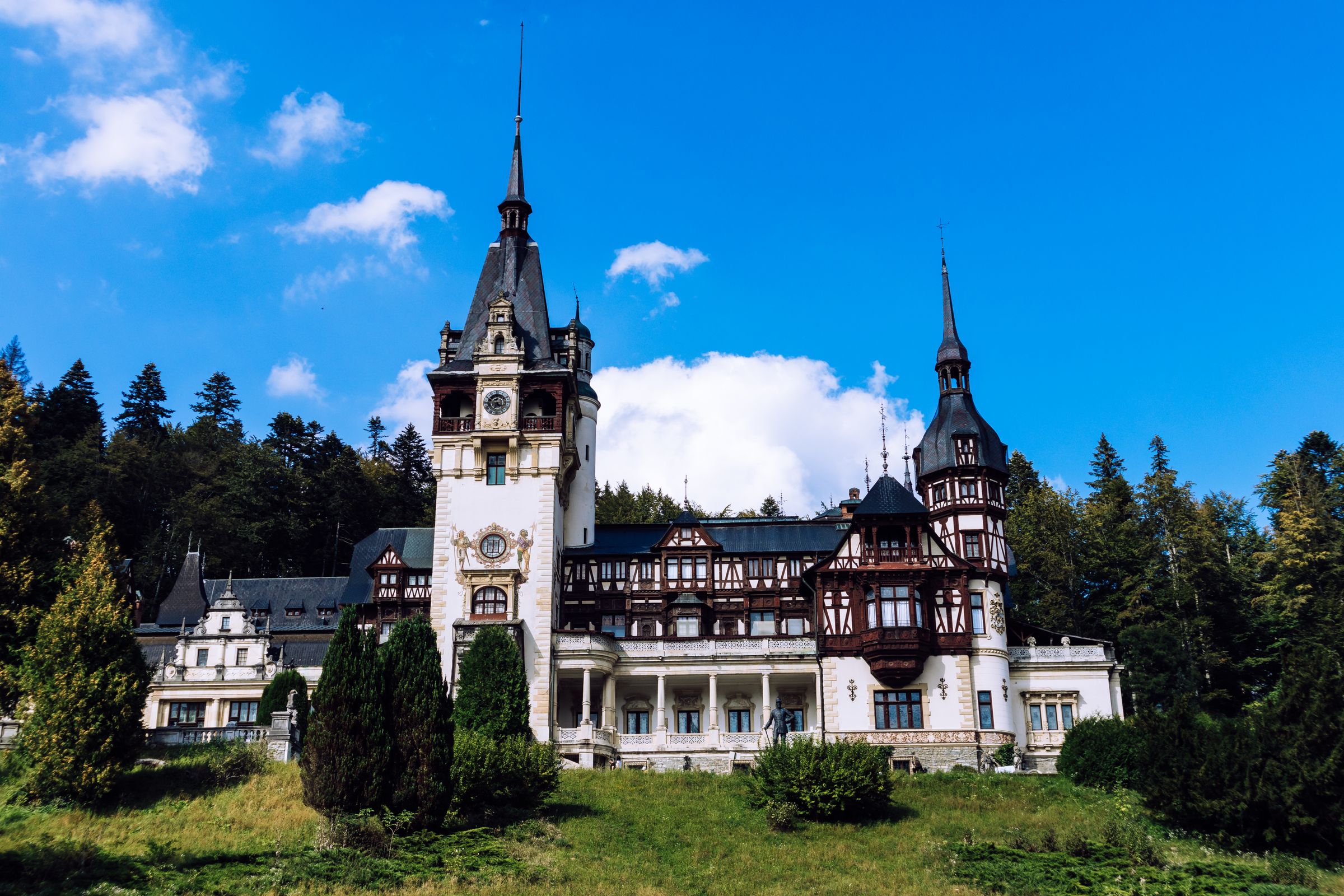 Peleș Castle's timbered towers and pale stone front above a grassy slope, with fir forest behind and a blue sky
