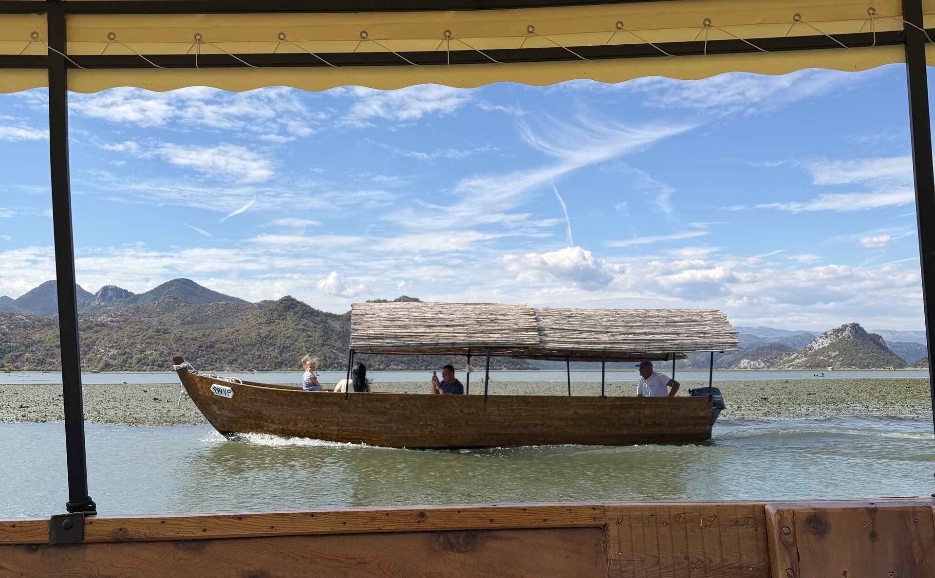 Another wooden tour boat with a reed roof crossing shallow water on Lake Skadar, seen from beneath the awning of a second boat