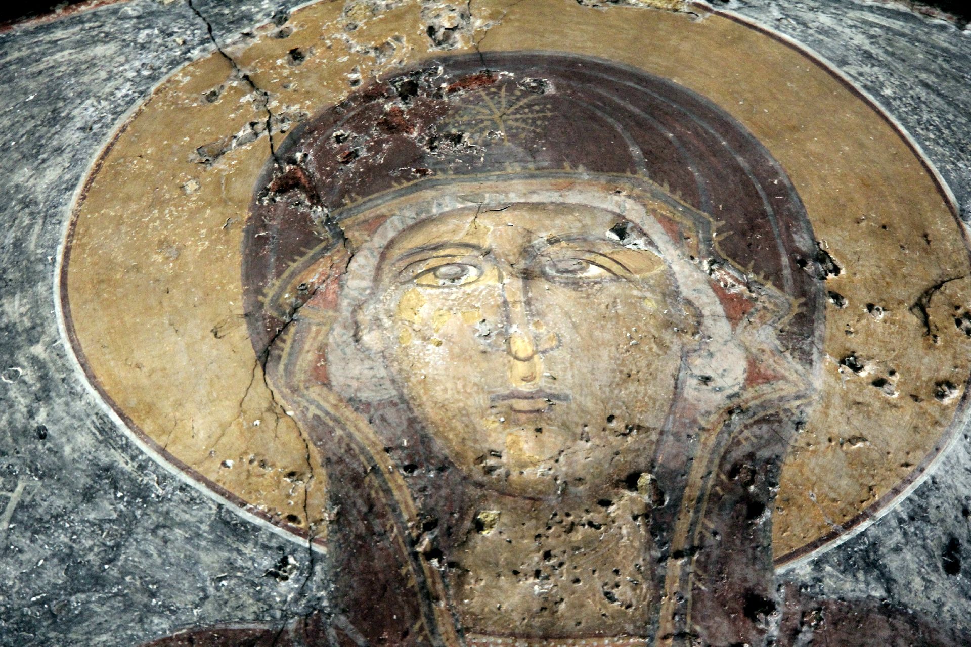 A damaged medieval fresco of a haloed figure in a dark maphorion, the plaster cracked and pitted