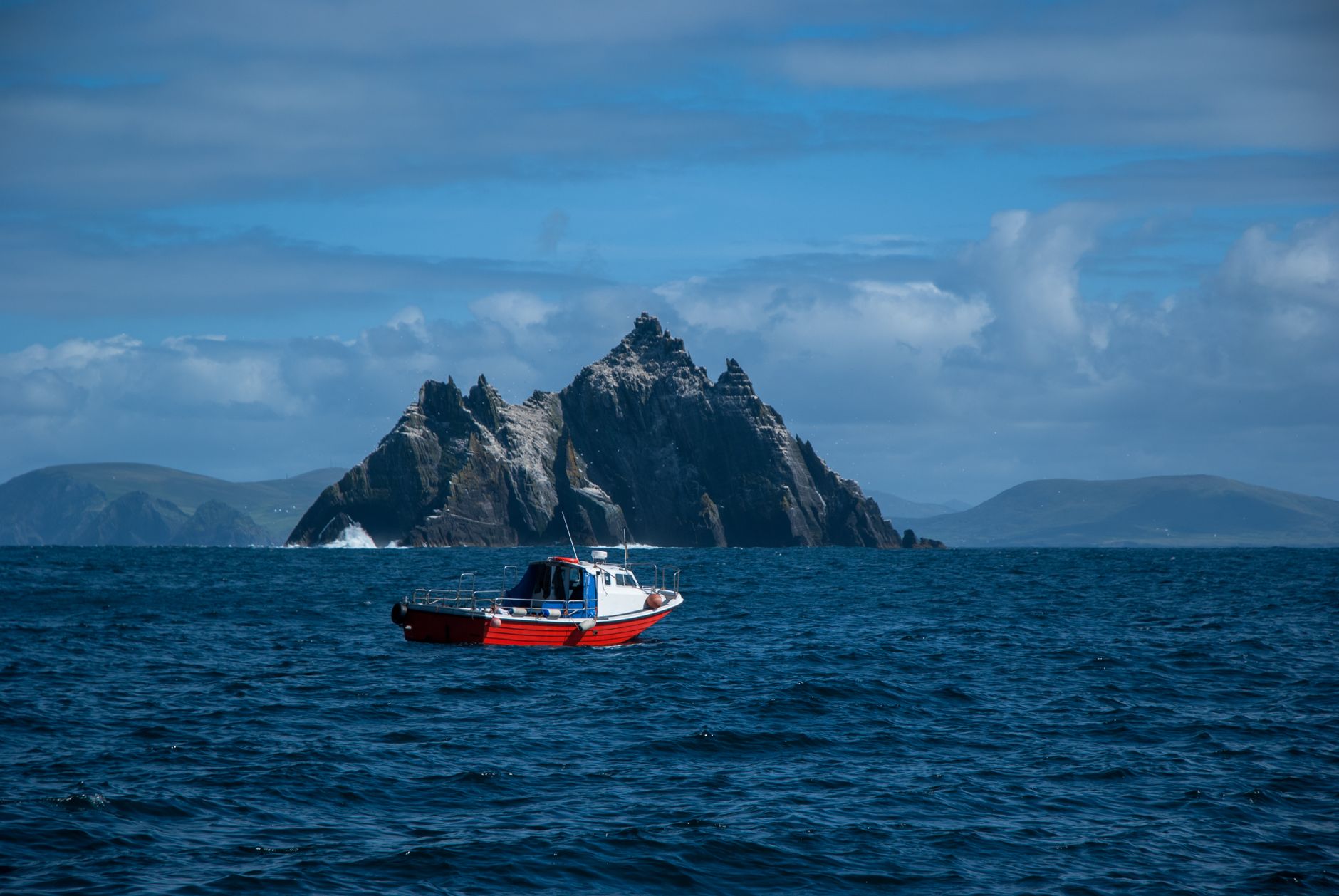 A small red and white boat on open Atlantic water beneath a jagged pinnacle island, the mainland faint behind