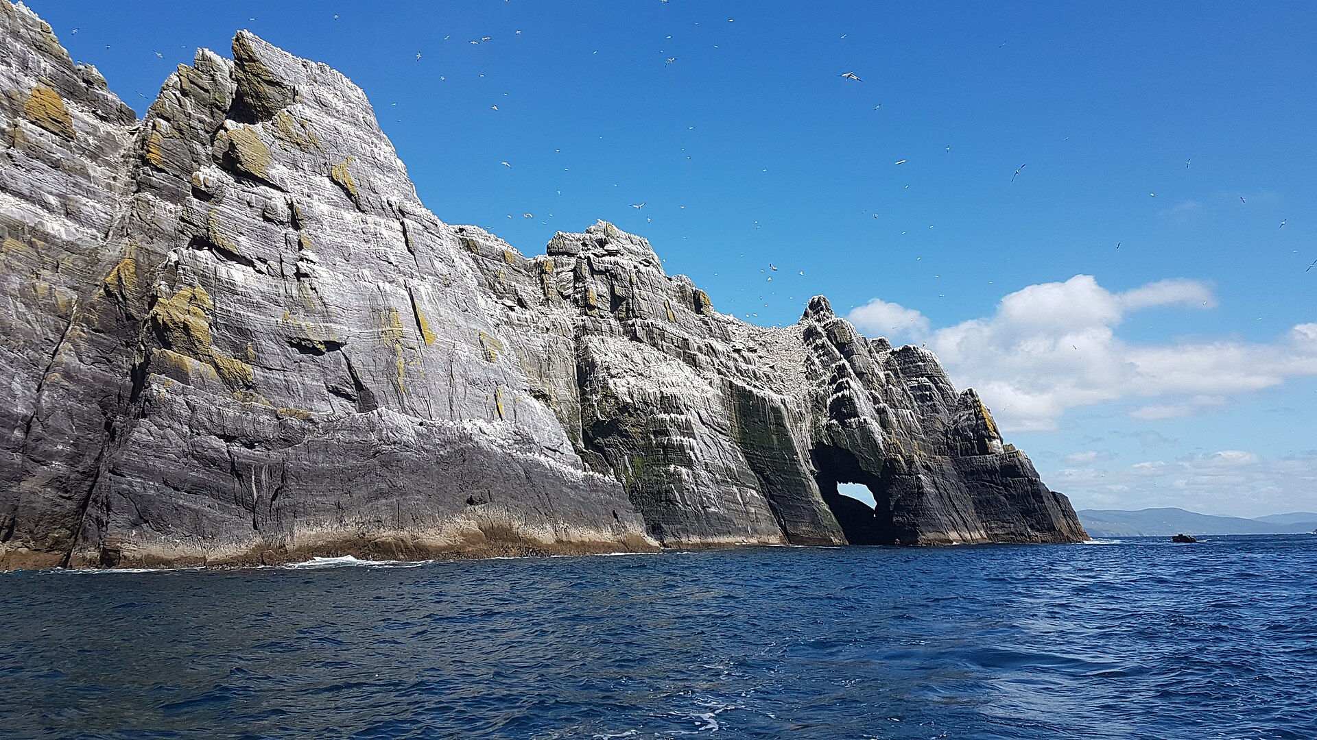 The white-streaked banded cliffs and sea arch of Little Skellig with gannets in the air around it